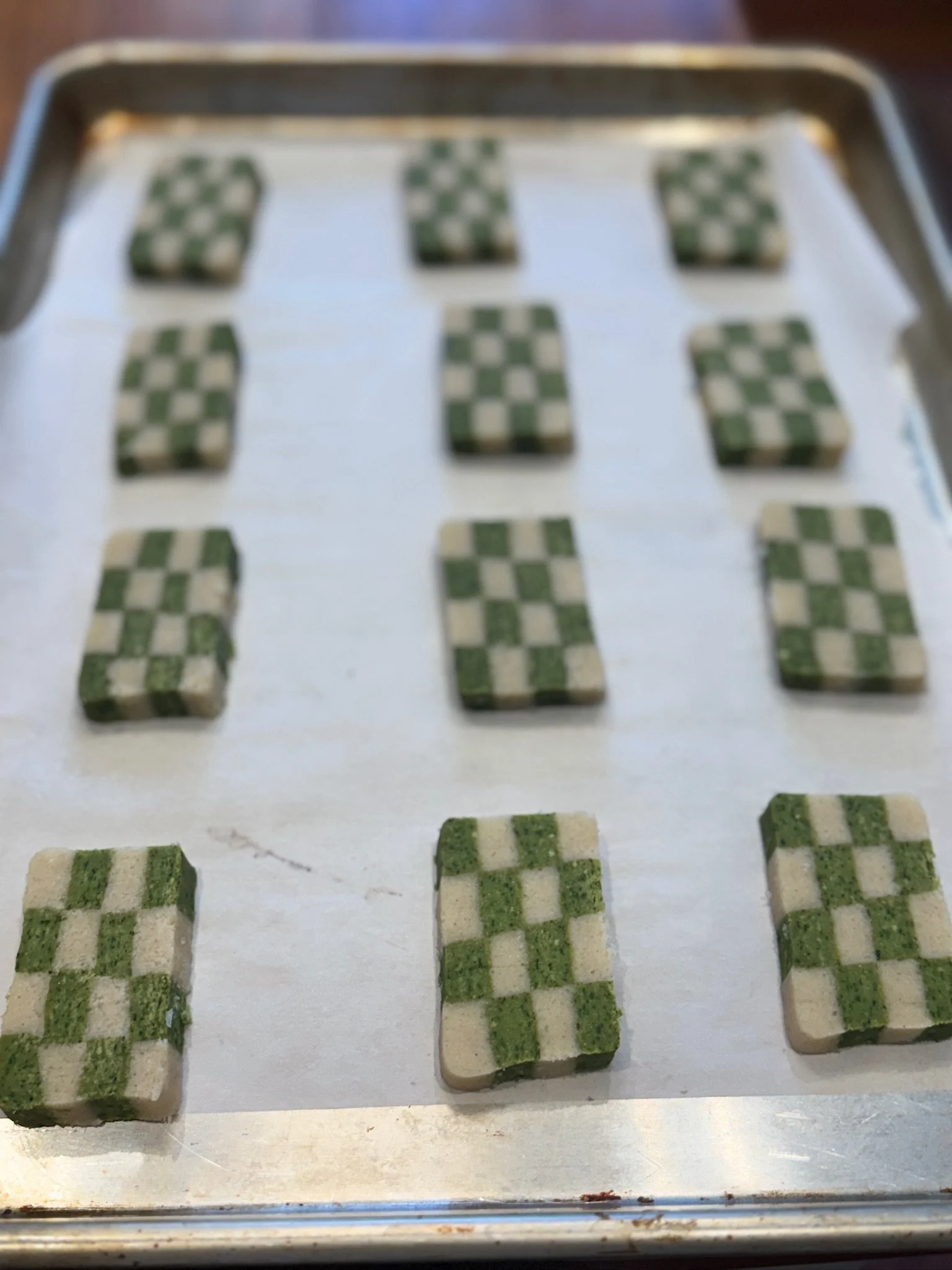 A baking sheet with multiple small, square green and white checkered basil parmesan shortbread cookies or candies arranged in rows.