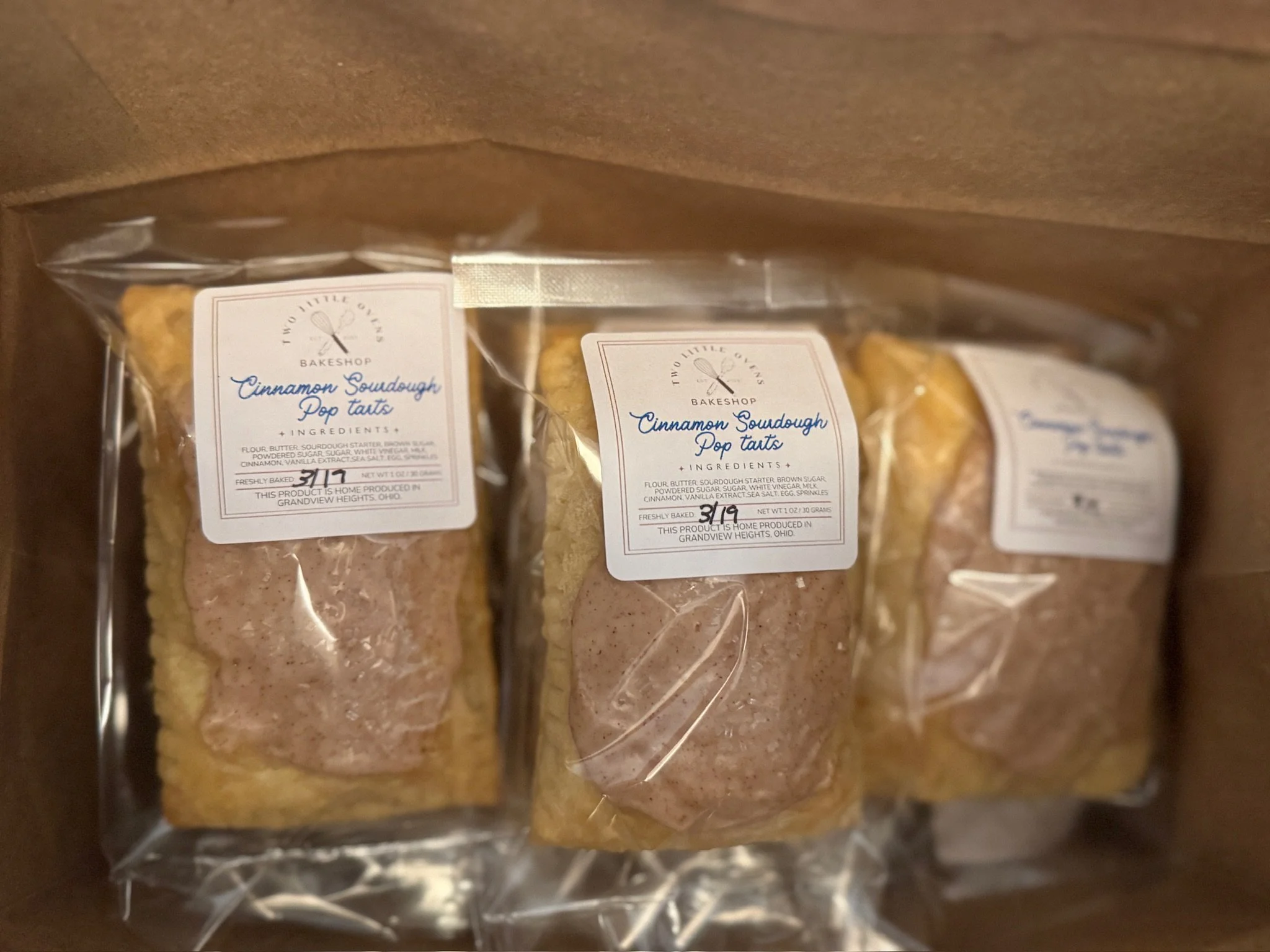 Three packages of cinnamon sourdough popcorn tarts in clear plastic packaging with labels from Two Little Ovens Bakeshop, produced in Ohio.