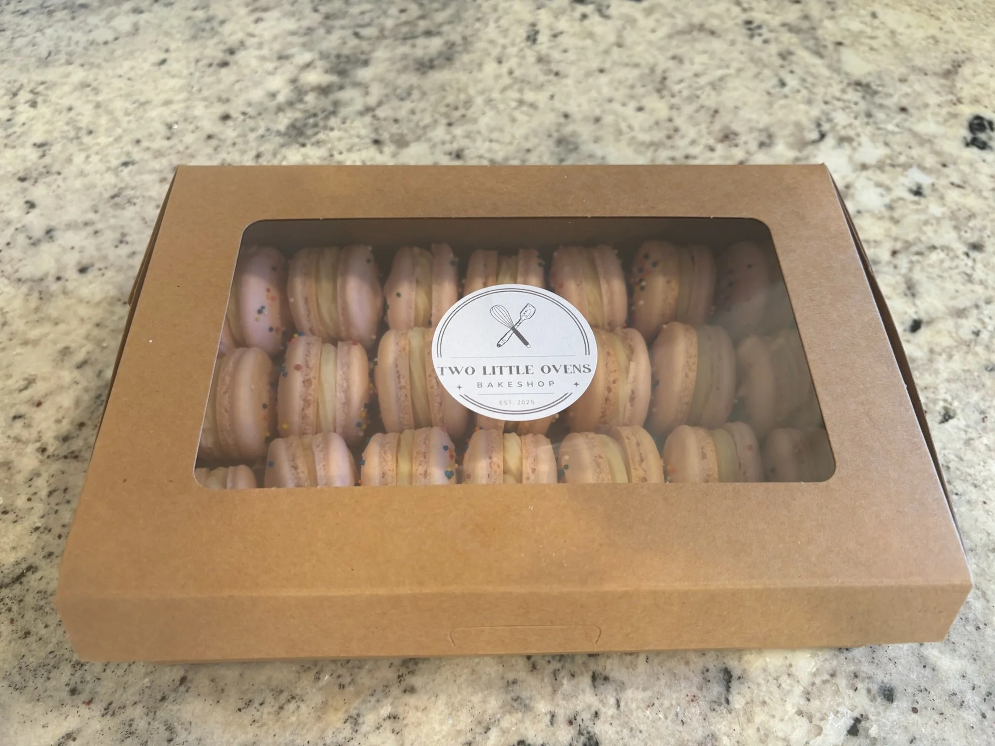 Box of pink macarons with colorful sprinkles from Two Little Ovens Bakeshop placed on a granite countertop.