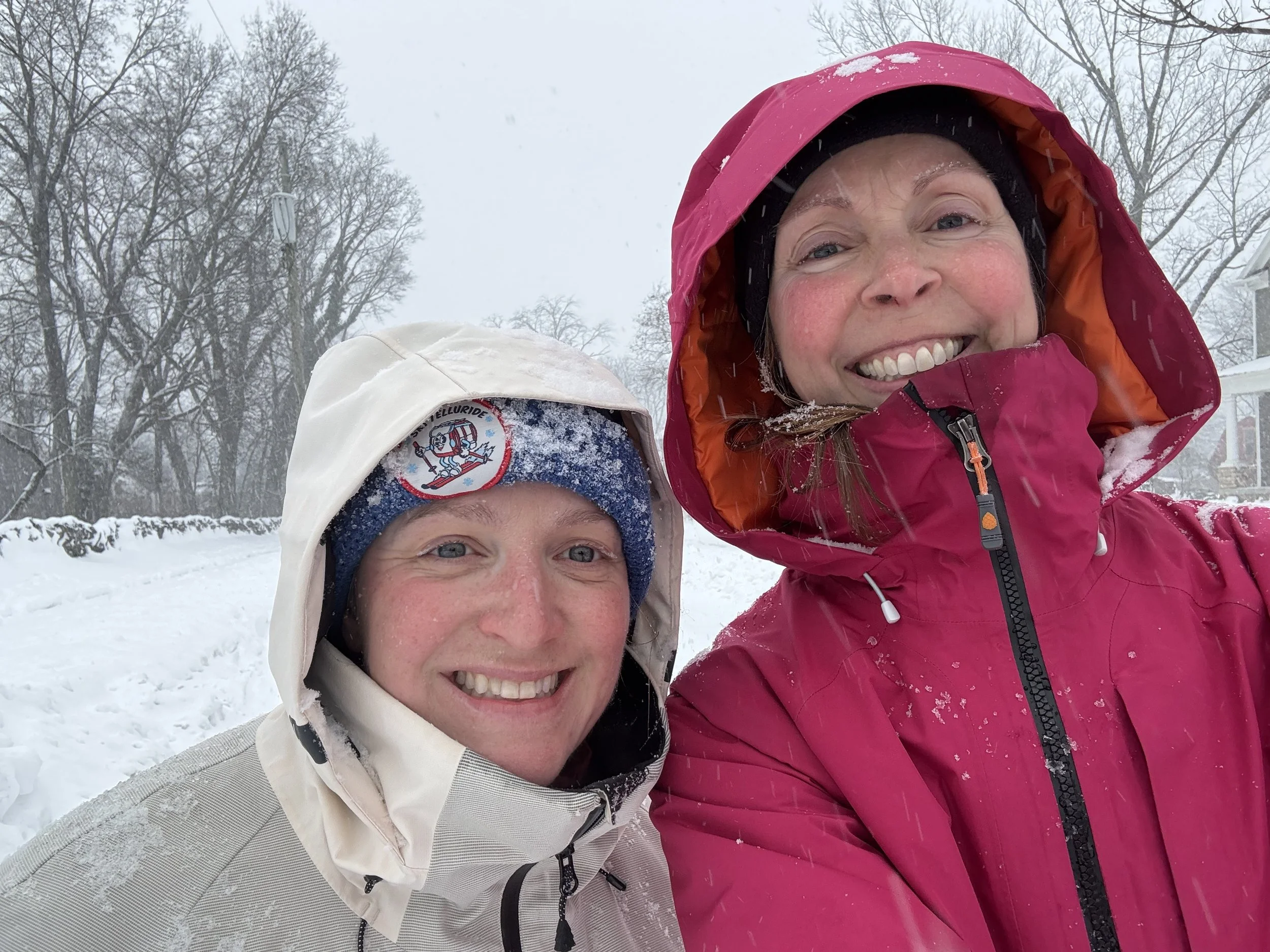 Two smiling women taking a selfie in a snowy outdoor setting, dressed warmly in winter jackets with snow on their clothing, with leafless trees in the background.