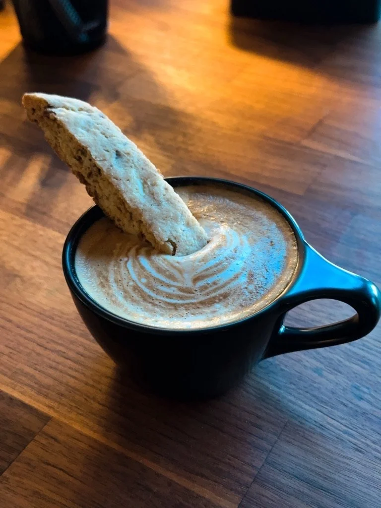 A black coffee mug filled with coffee topped with foam, garnished with a large biscotti cookie.