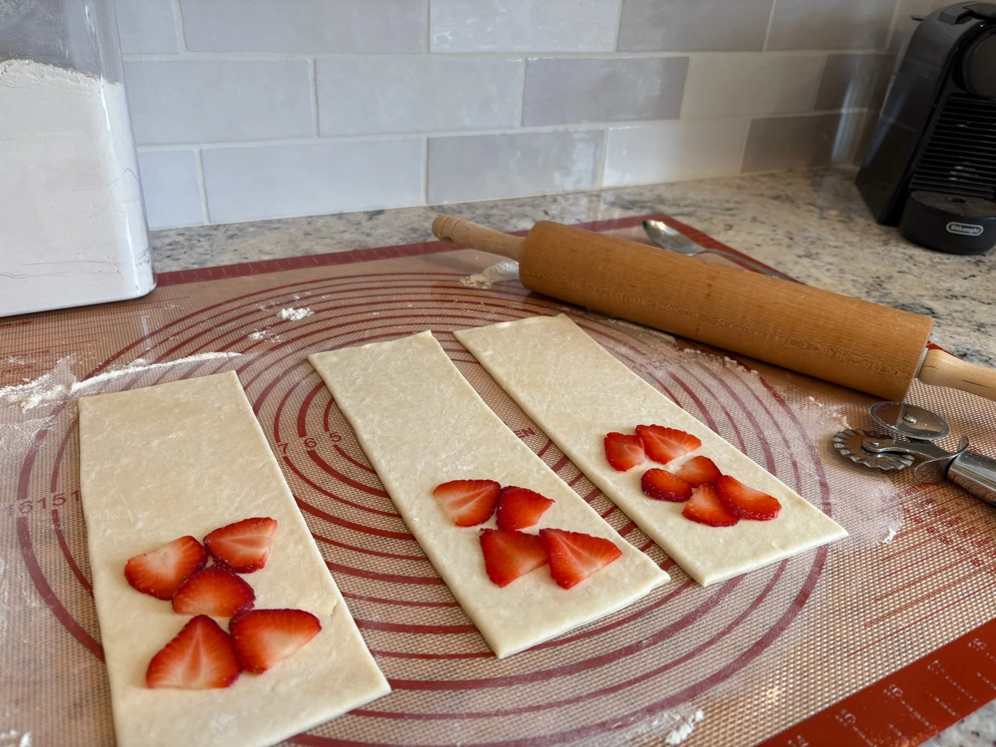 Slices of puff pastry topped with sliced strawberries, on a silicone baking mat. A rolling pin and a spoon are nearby on the kitchen counter.