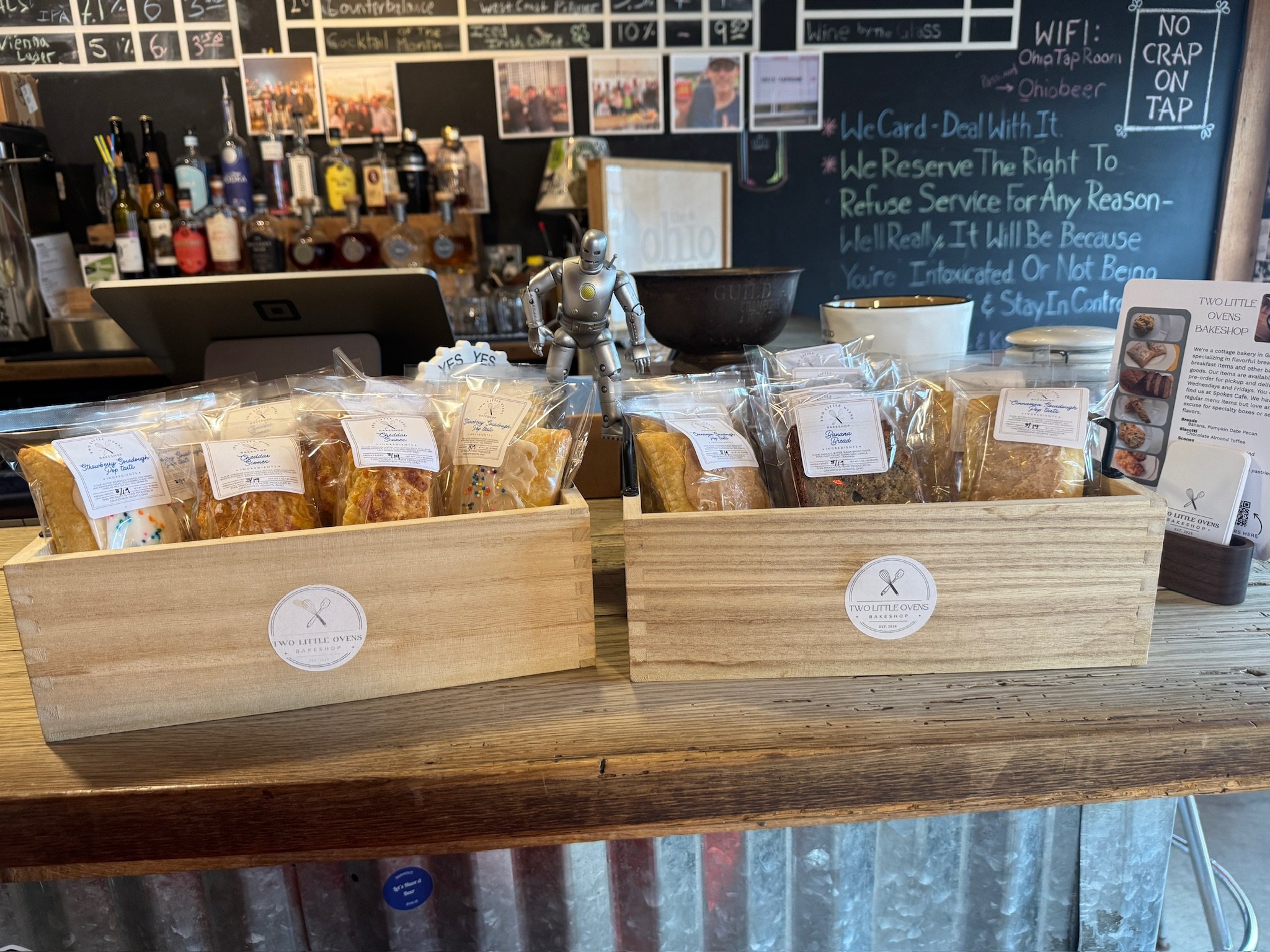 A display of assorted baked goods in clear plastic bags in a bakery or cafe setting with a chalkboard menu in the background.