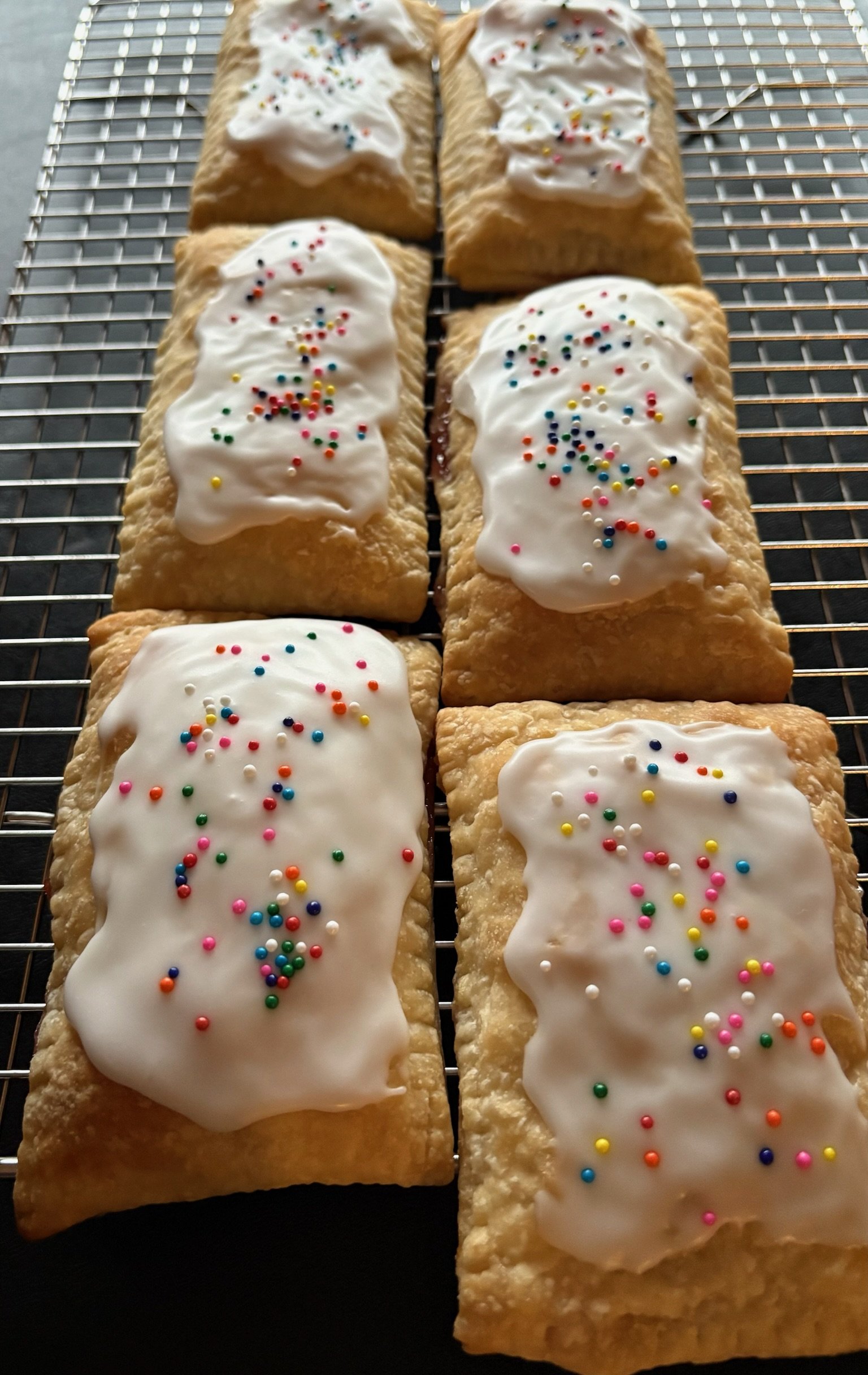 Six rectangular strawberry pastries with white icing and colorful sprinkles on top, arranged on a cooling rack.