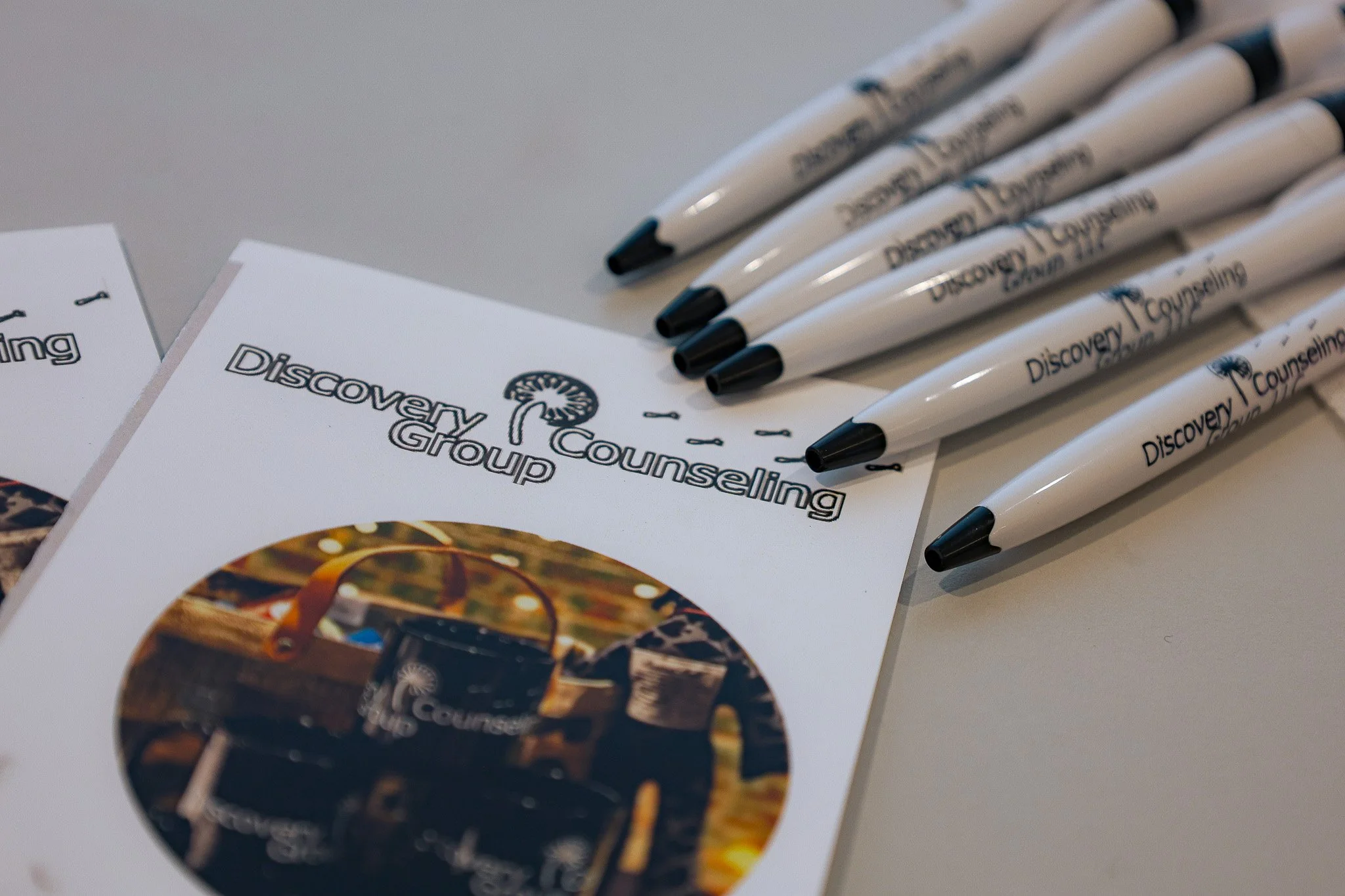 A stack of white promotional pens with black tips and clip, laying beside printed flyers for Discovery Counseling, with a circular photo of a cityscape or art installation on the flyer.