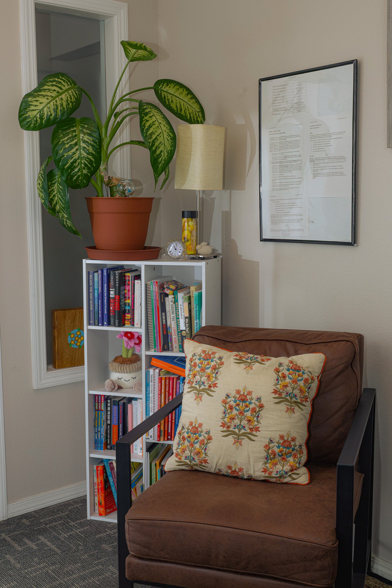 A cozy reading corner featuring a brown armchair with a decorative embroidered pillow, a white bookshelf filled with books, a tall indoor plant, a table lamp, and a framed document on the wall.