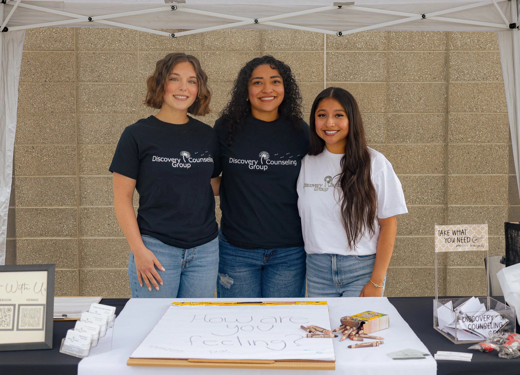 Three young women standing together under a white canopy over a table at an outdoor event. They are smiling and wearing black and white T-shirts that read "Discovery Counseling Group." The table in front of them has signs, brochures, and writing supplies, with a large paper displaying the question "How are you feeling?" written on it.
