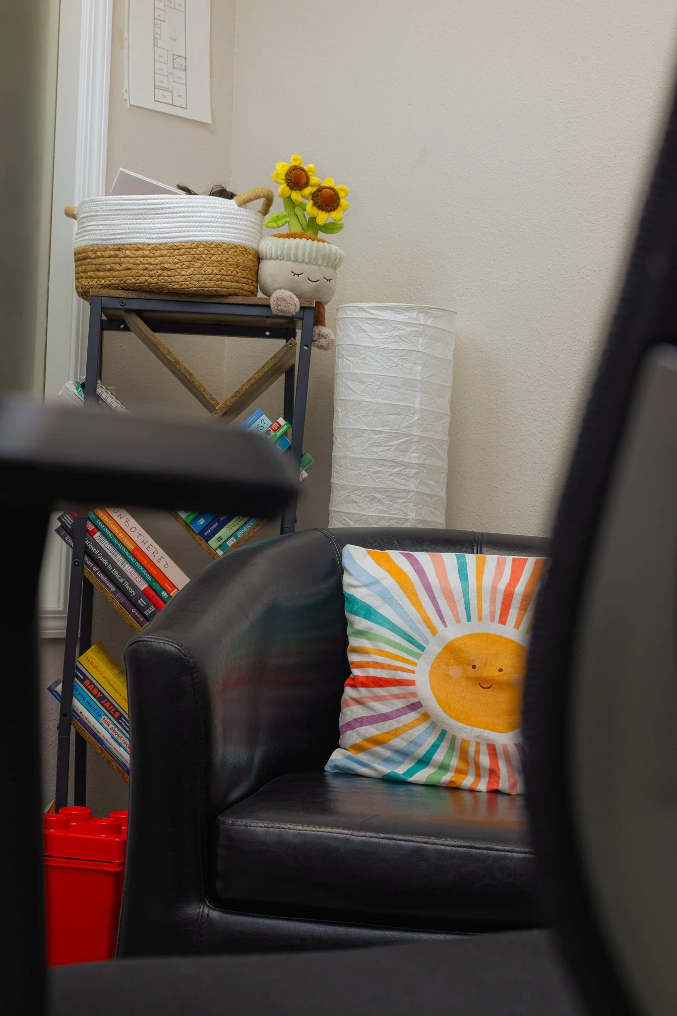 A black leather armchair with a colorful striped pillow featuring a smiling sun design. Behind the chair is a white paper lantern, a metal shelf with books and a plush toy with yellow flowers on top, and a framed diagram on the wall.