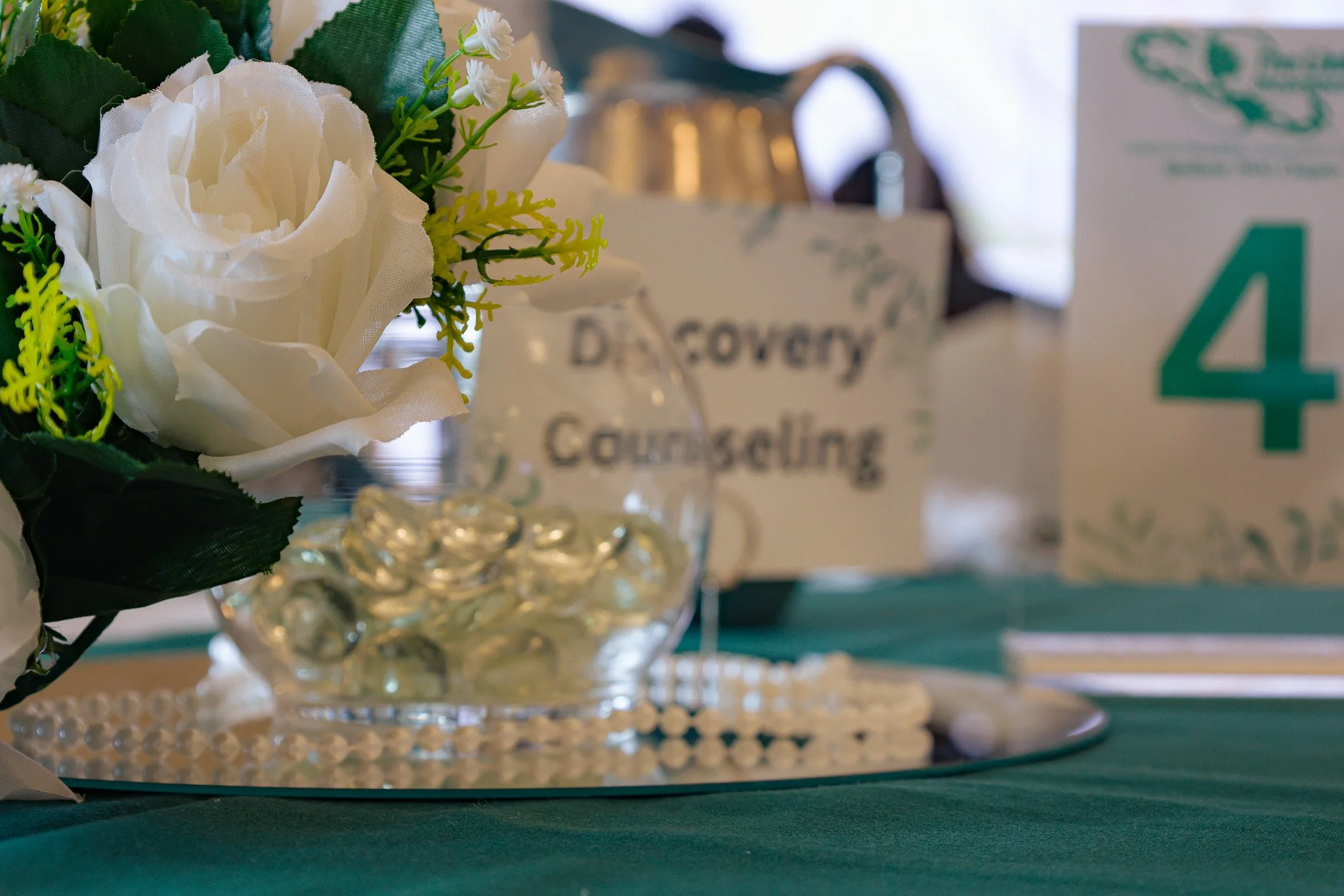 A floral arrangement with white artificial flowers and greenery, placed on a decorative glass and pearl tray, in front of a blurred background featuring signs with the number 4 and the word 'County'.