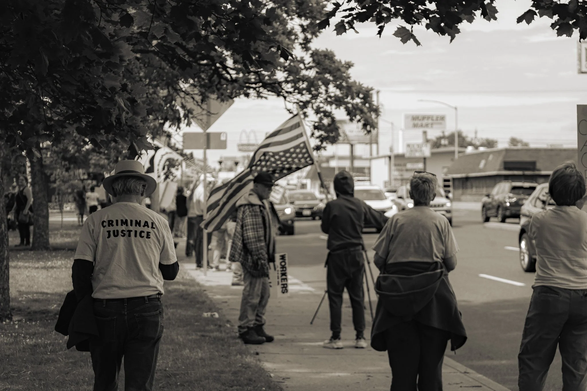 People participating in a protest march, some holding American flags, with one person wearing a jacket labeled 'Criminal Justice'. The scene is in a city street with cars and buildings in the background, and trees lining the sidewalk.