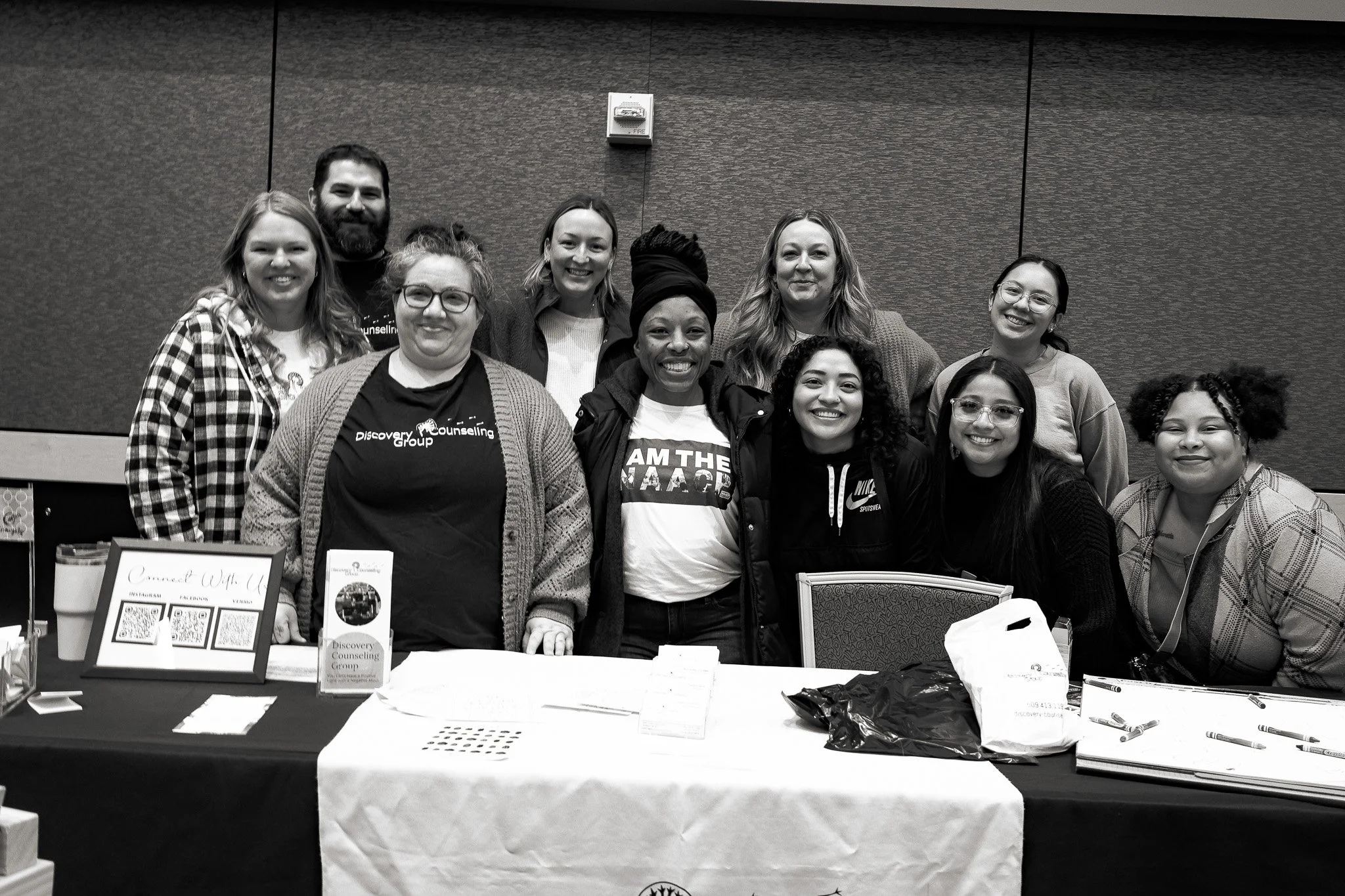 Group of diverse women and one man smiling at a table with promotional materials, QR codes, and pens at an indoor event or conference.