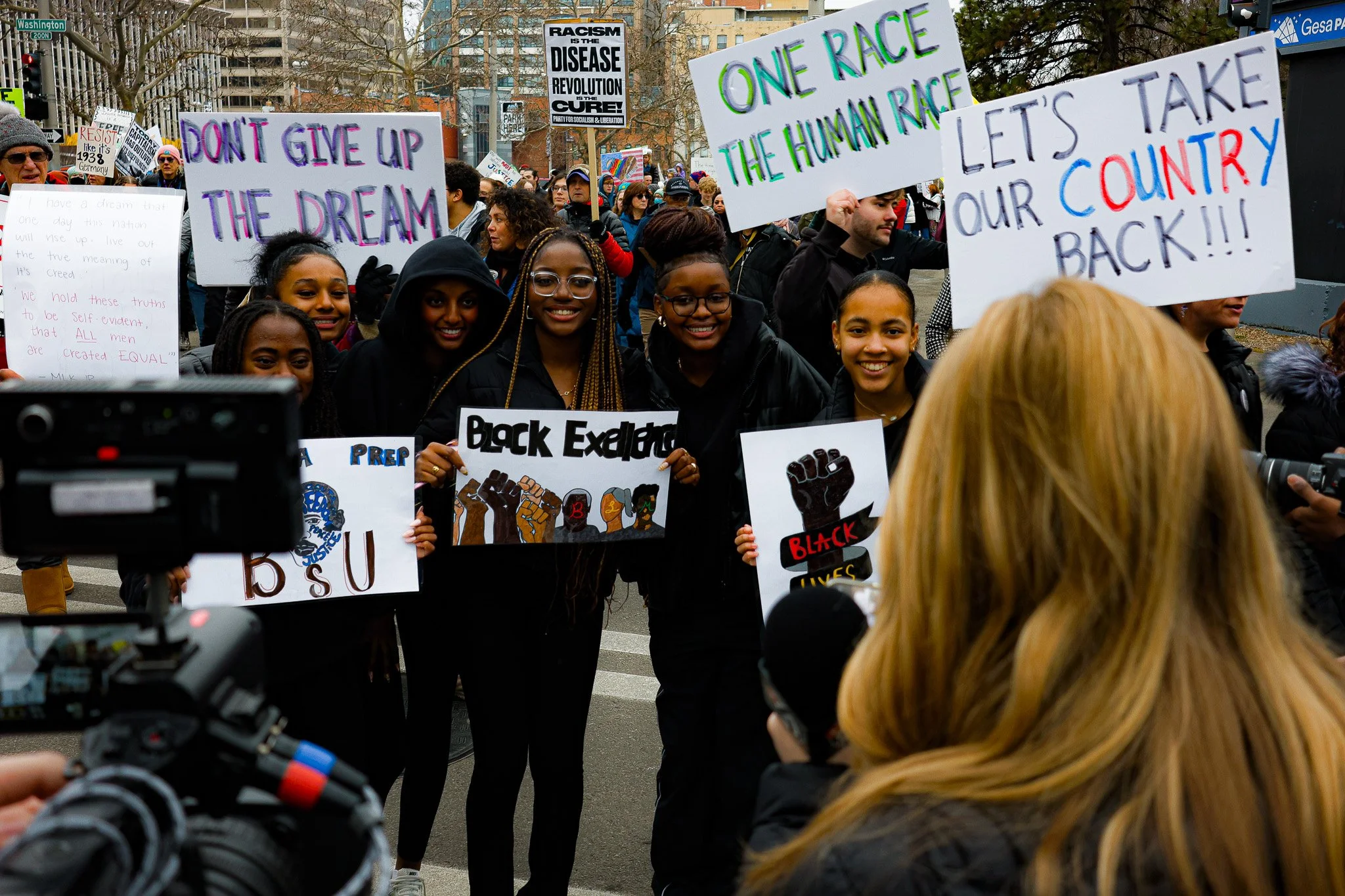 A diverse group of protesters holding signs with messages about racial justice, including slogans like 'Don't give up the dream,' 'One race, the human race,' and 'Let's take our country back.' The crowd appears to be participating in a demonstration or rally on a city street.
