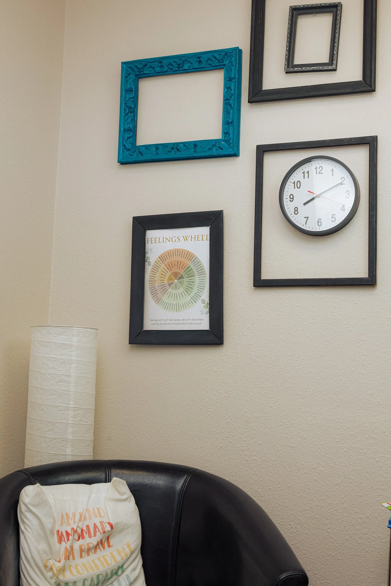 A wall decorated with five picture frames of various sizes and colors, a clock, and a framed feelings wheel. Part of a black leather chair and a white pillow with orange text are visible in the foreground, and a white paper lamp is on the left.