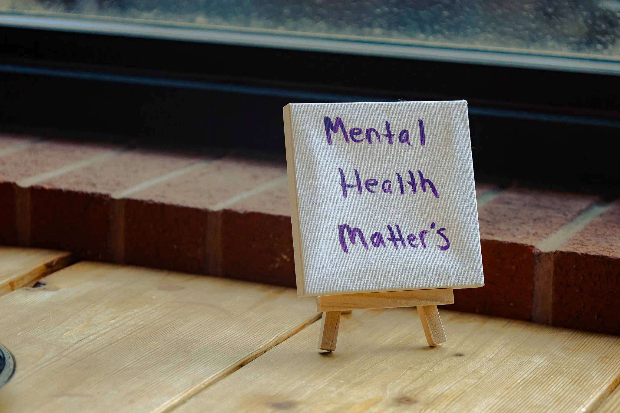 A small easel holding a white card with handwritten purple text that reads 'Mental Health Matters'. The easel is placed on a wooden surface with a brick wall behind it.