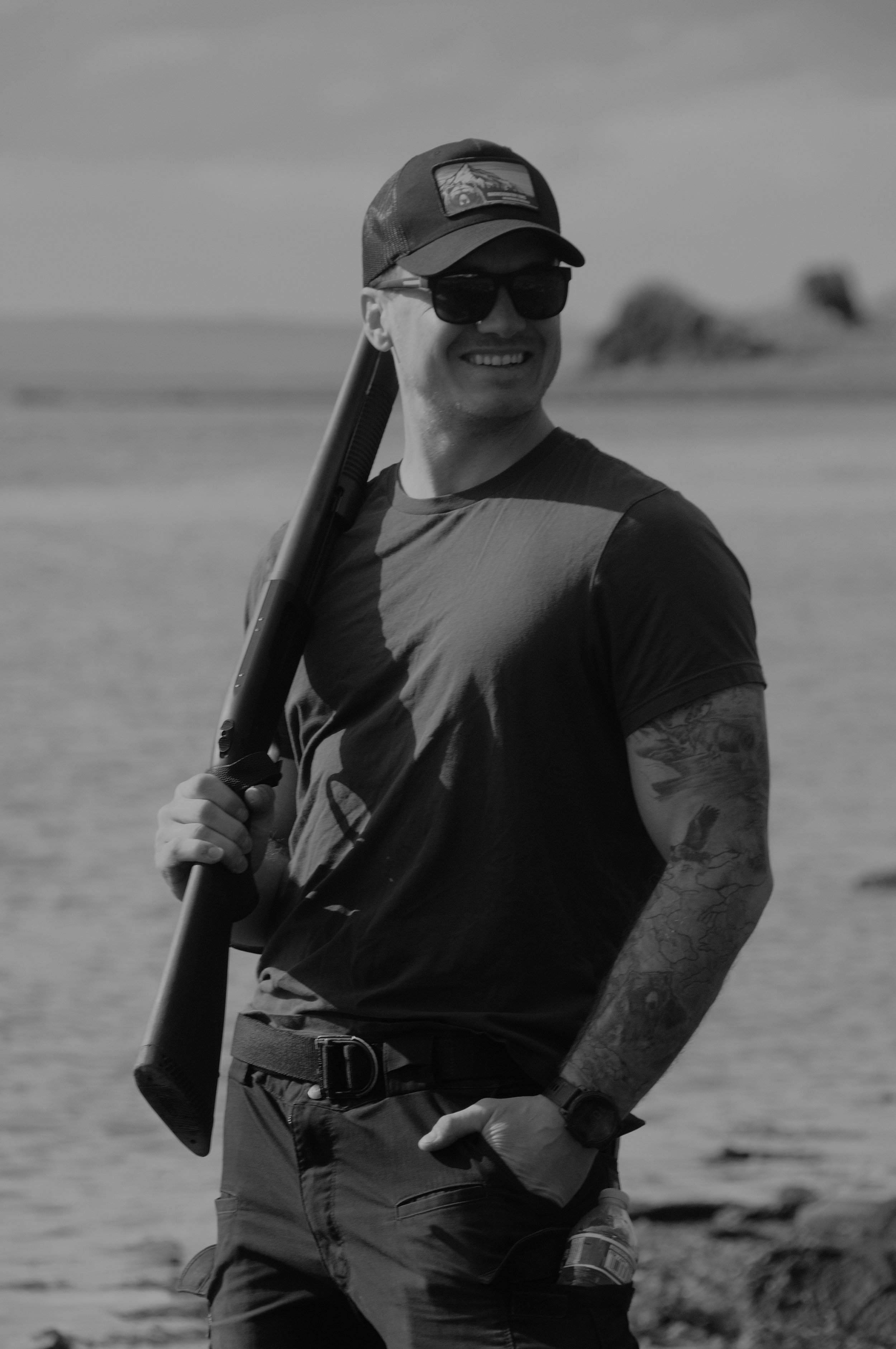 A man smiling and walking outdoors on a beach, wearing sunglasses, a cap, a t-shirt, and carrying a water bottle and a stick, with a blurred background of sand and rocks.