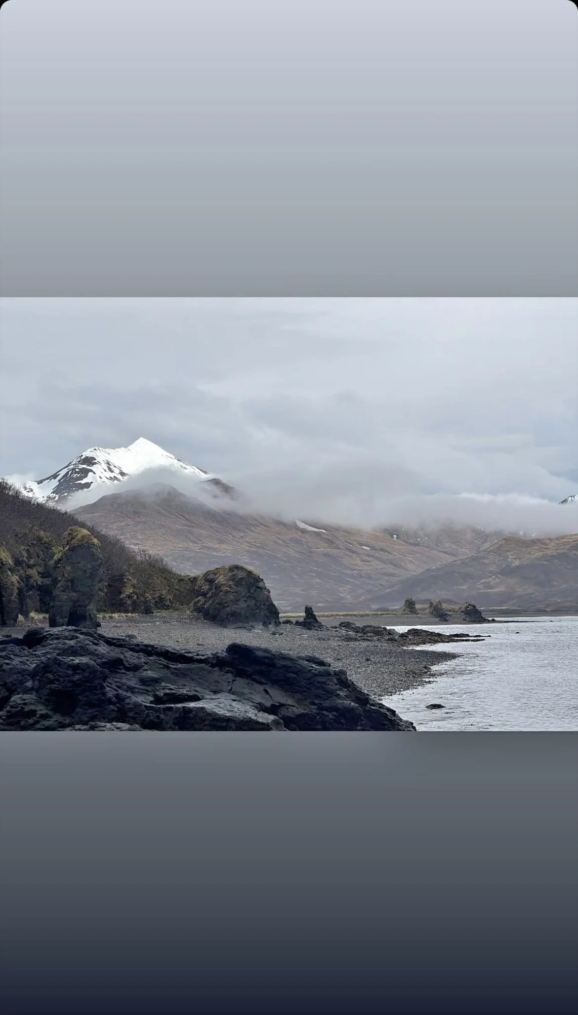 Scenic view of a snow-capped mountain with clouds, rocky shoreline, and body of water in the foreground.