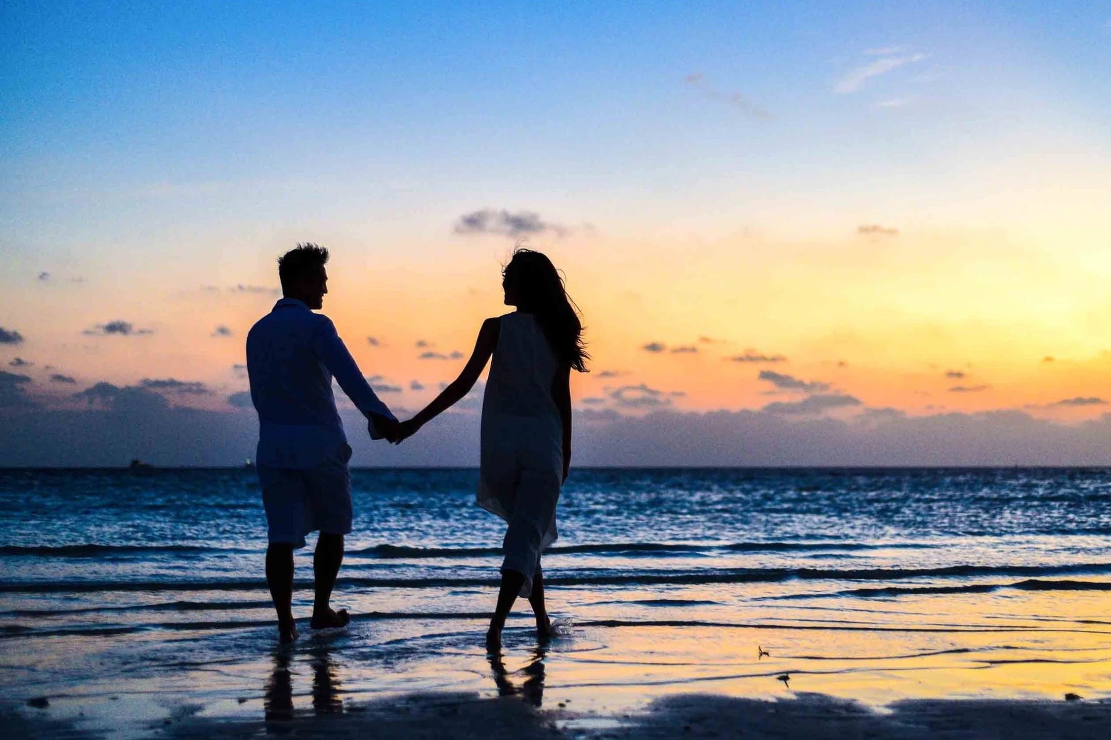 A silhouette of a man and woman holding hands and facing each other on the beach during sunset, with ocean waves and a colorful sky in the background.