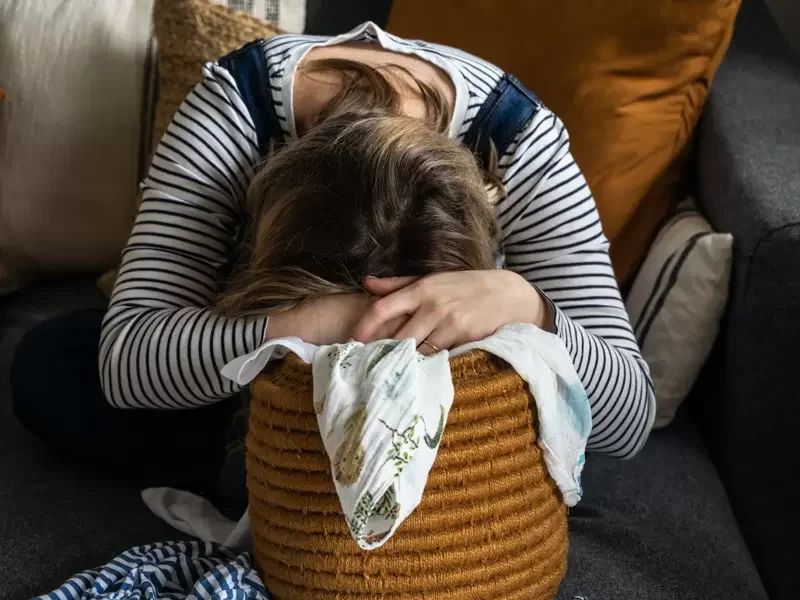 Person sitting on a sofa with their head resting on their arms, which are folded over a large, brown, textured woven basket in a cozy living room.