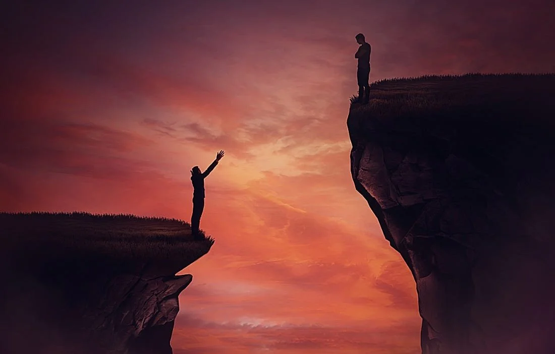 A person standing on a small grassy cliff reaching out to another person standing on a larger rocky cliff, with a sunset sky in the background.