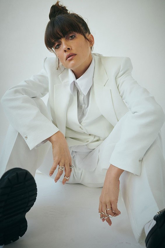 Woman with dark hair and bangs wearing a white suit, sitting on the floor against a plain white background.