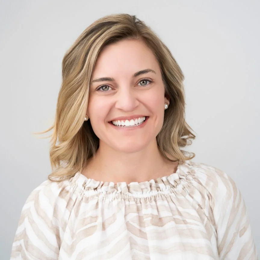 A smiling woman with blonde hair in a loose wave, wearing pearl earrings and a cream-colored striped blouse, against a plain light gray background.