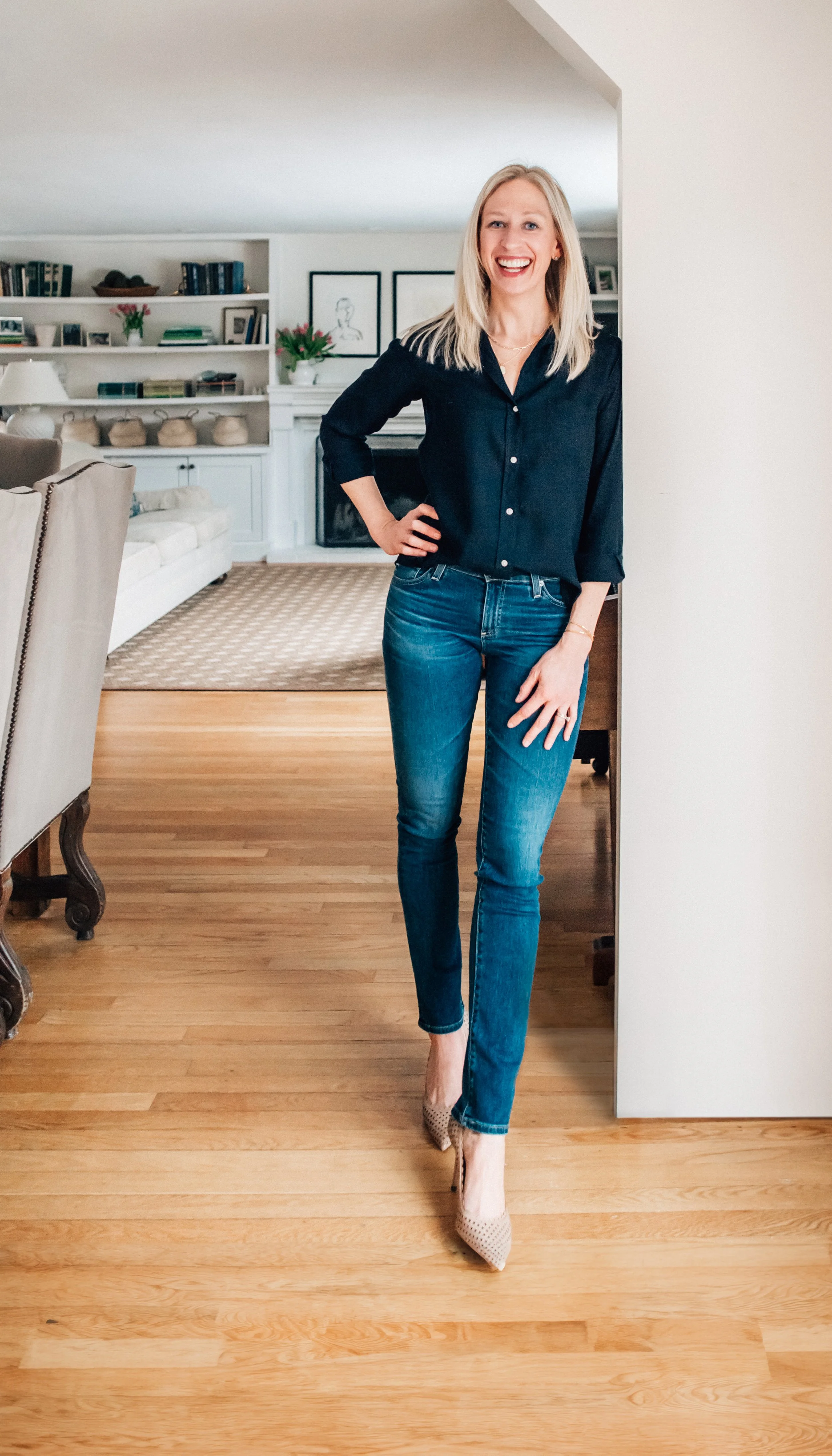 A woman with blonde hair standing in a living room, smiling, wearing a black shirt, blue jeans, and beige high heels.