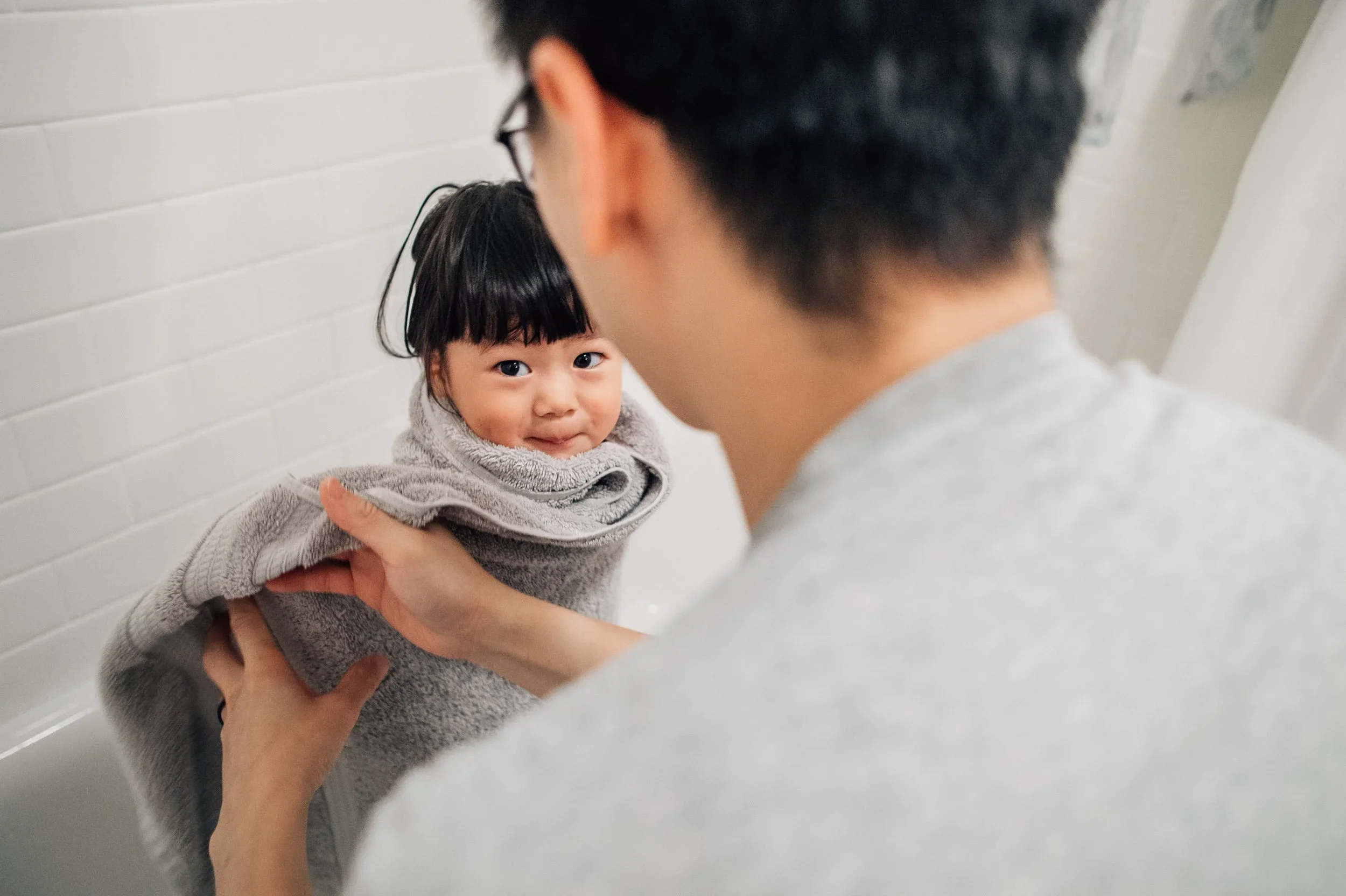 A young girl with short dark hair and bangs is being held up by an adult, facing each other in a room with white tiled walls. The girl looks directly at the camera with a mild smile.