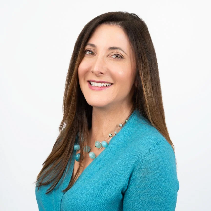 Portrait of a woman with long brown hair, smiling, wearing a turquoise top and matching necklace, against a white background.