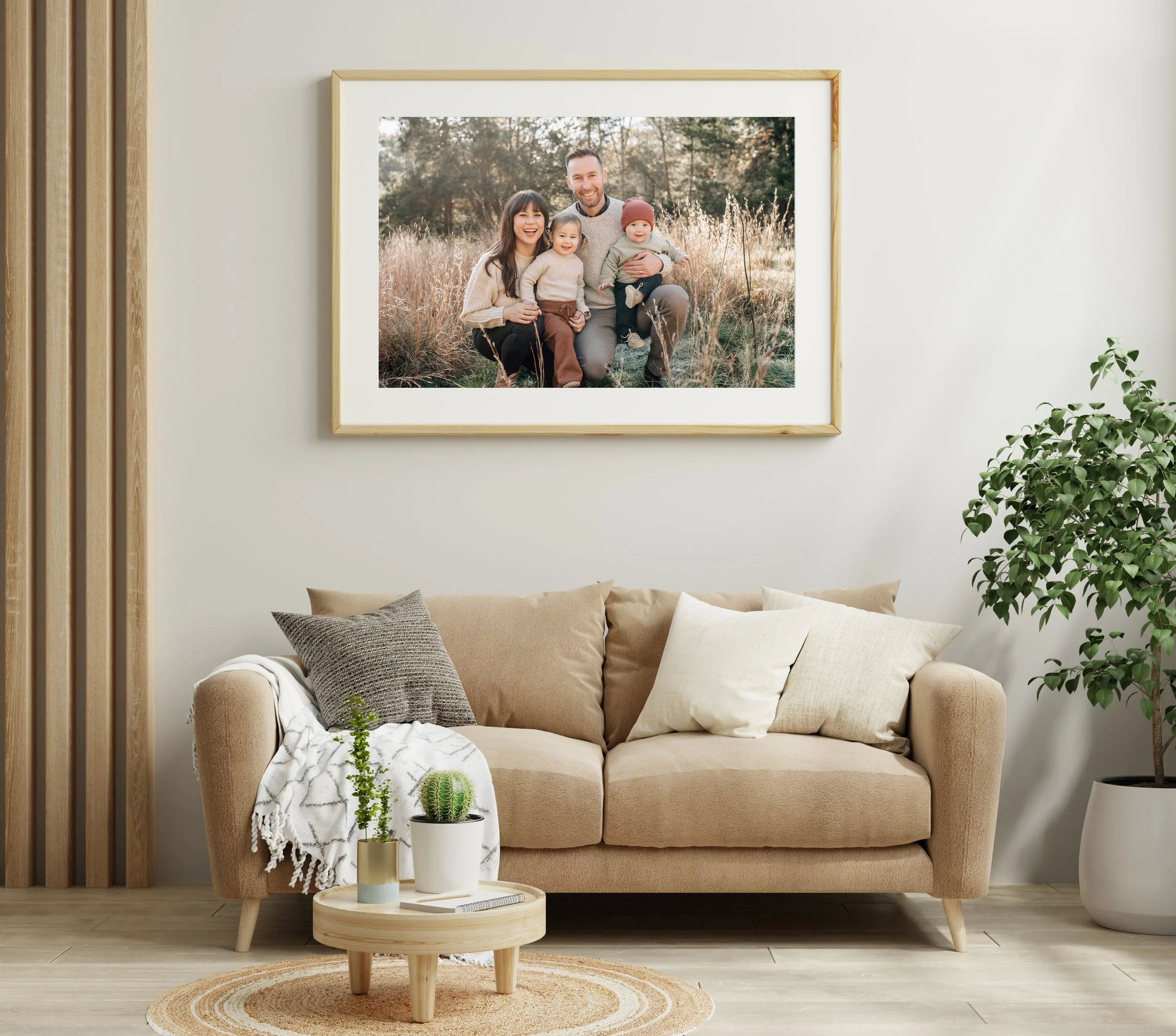 Living room with a beige sofa, a small round wooden coffee table with potted cacti, throw pillows, a patterned throw blanket, a framed family photo on the wall, and potted plants.
