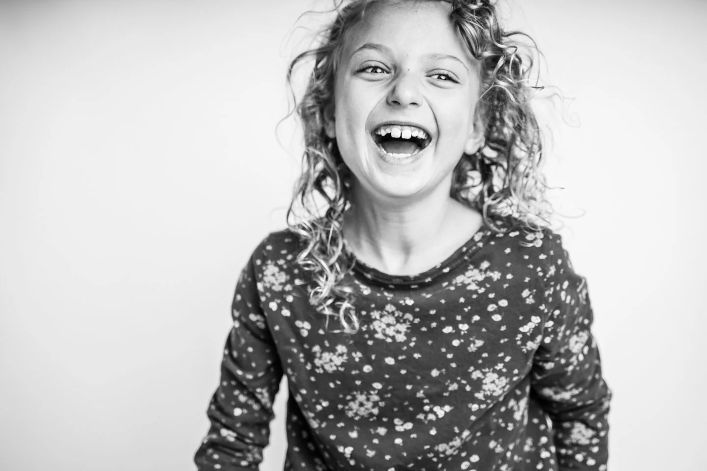 A young girl with curly hair laughing joyfully, wearing a dark shirt with a floral pattern.