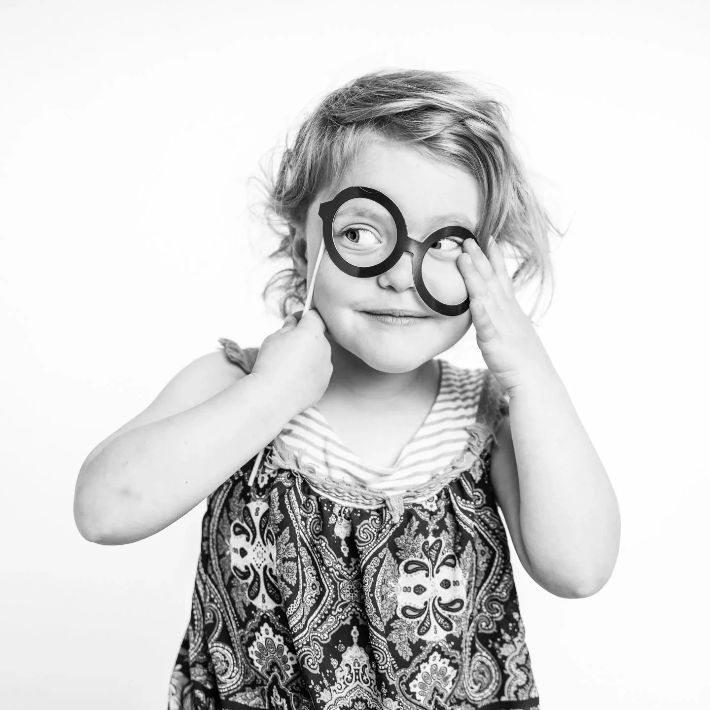 A young girl with curly hair wearing a patterned dress, holding novelty glasses with large round frames and a stick, looking to her left with a playful expression.