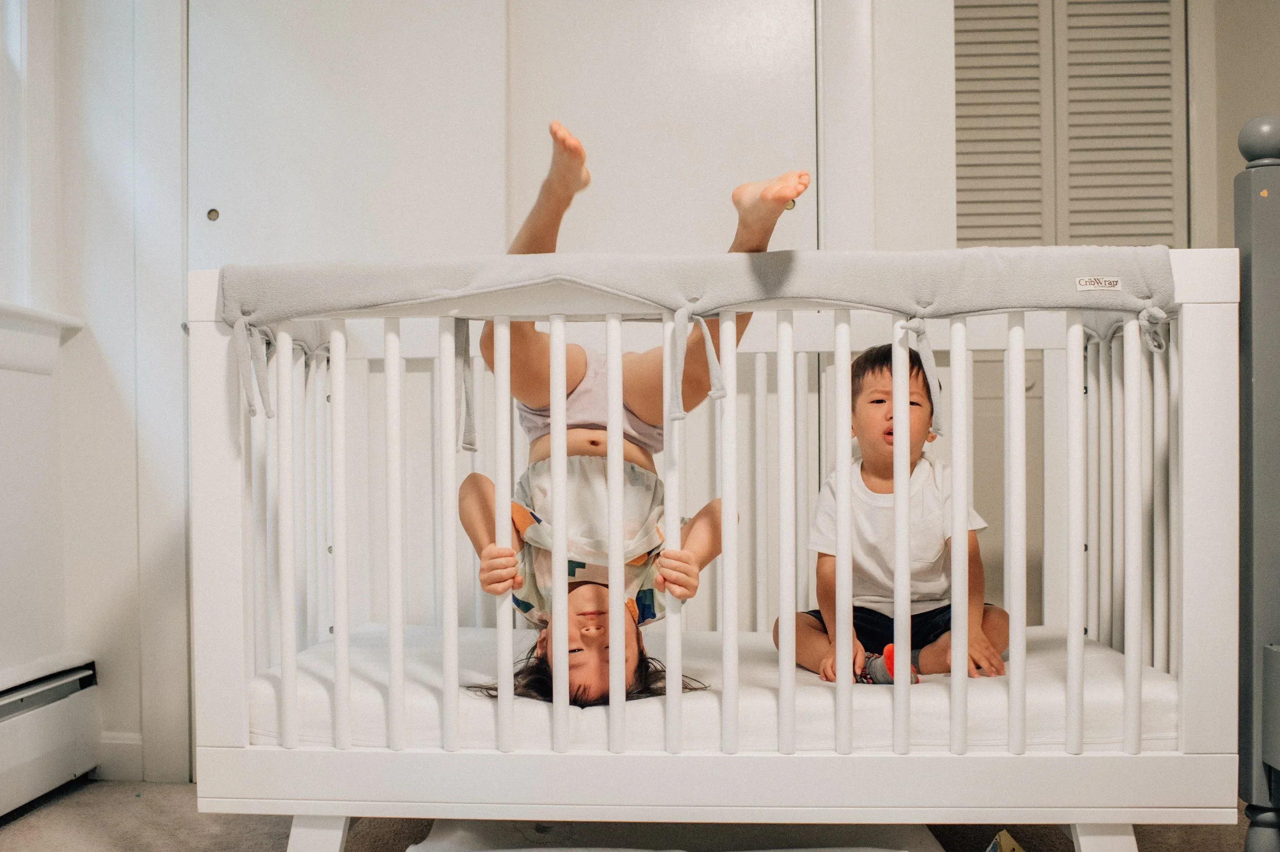 Three young children are inside a white crib with bars, two are hanging upside down and one is sitting upright. The crib is in a room with pale-colored walls and a closed door or closet behind it.