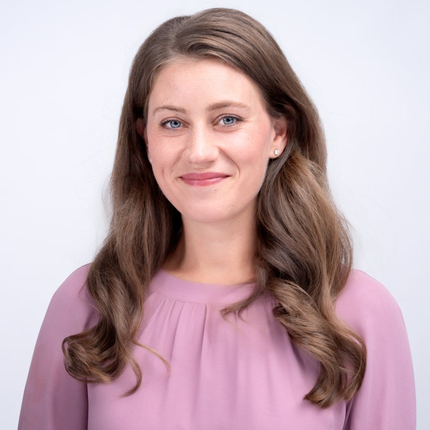 A young woman with long, wavy brown hair and blue eyes smiling at the camera, wearing a light purple blouse, standing against a plain white background.