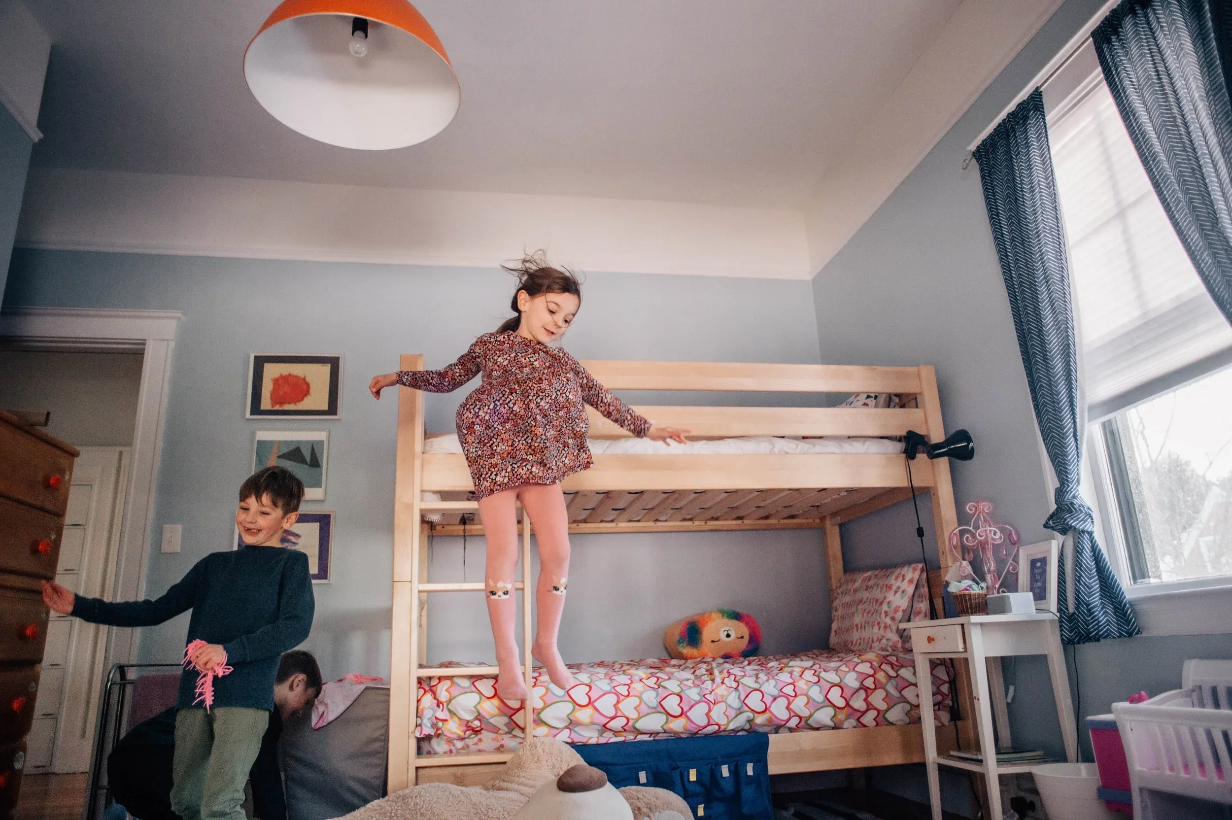 Children playing in a bedroom with a bunk bed. A girl stands on the top bunk, and two boys are on the lower level.