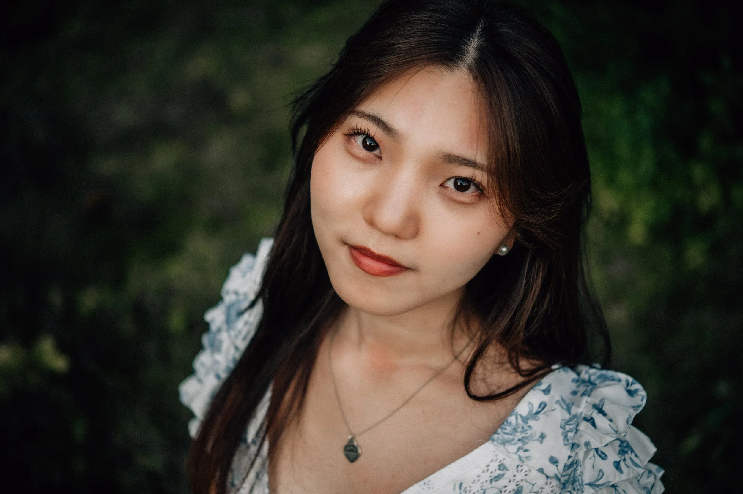 Close-up of a young woman with long dark hair, wearing a patterned blouse and a necklace, standing outdoors with blurred greenery in the background.
