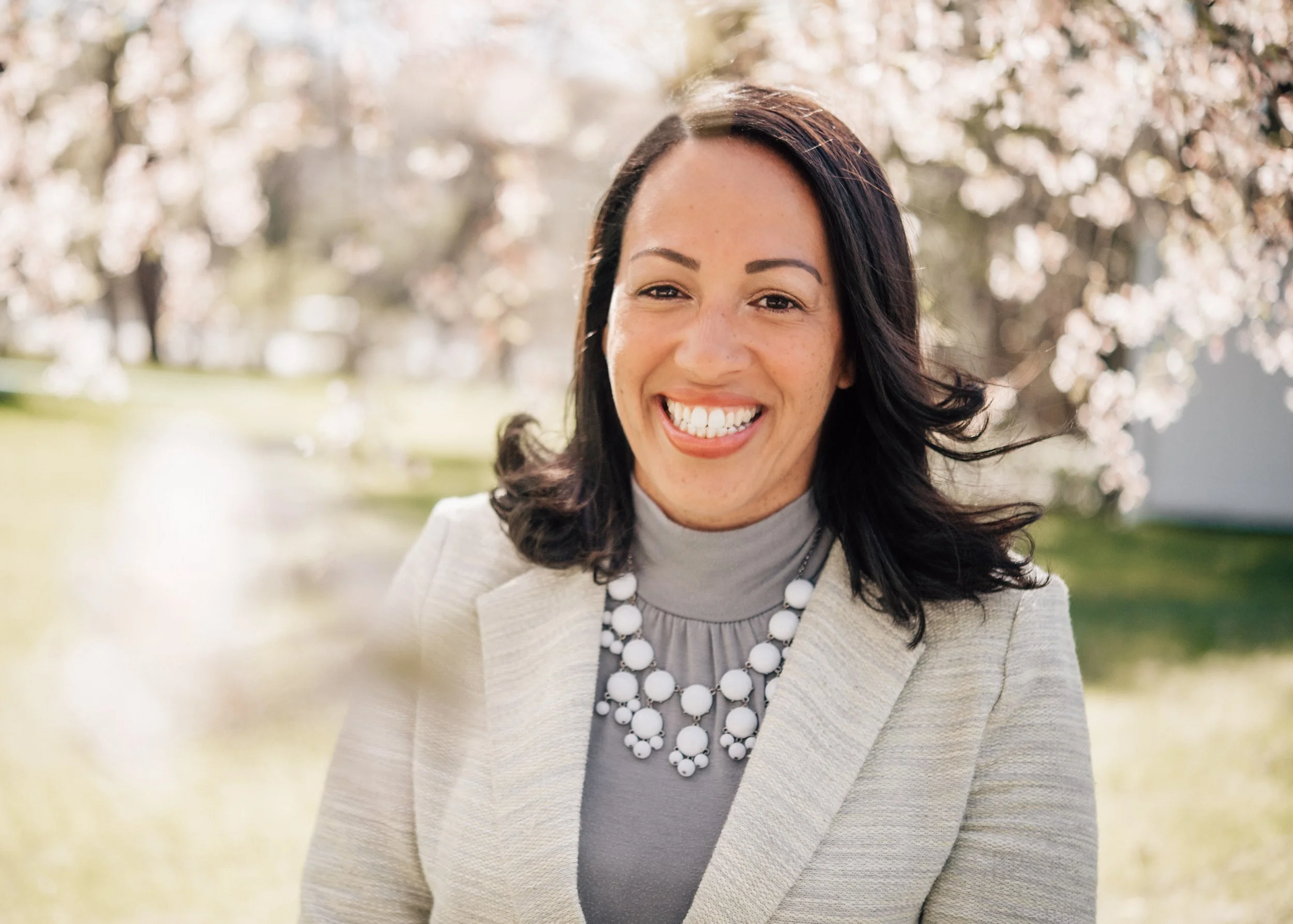 A smiling woman with dark wavy hair, wearing a light-colored blazer, gray turtleneck, and a large white bead necklace, standing outdoors with blooming cherry blossom trees in the background.