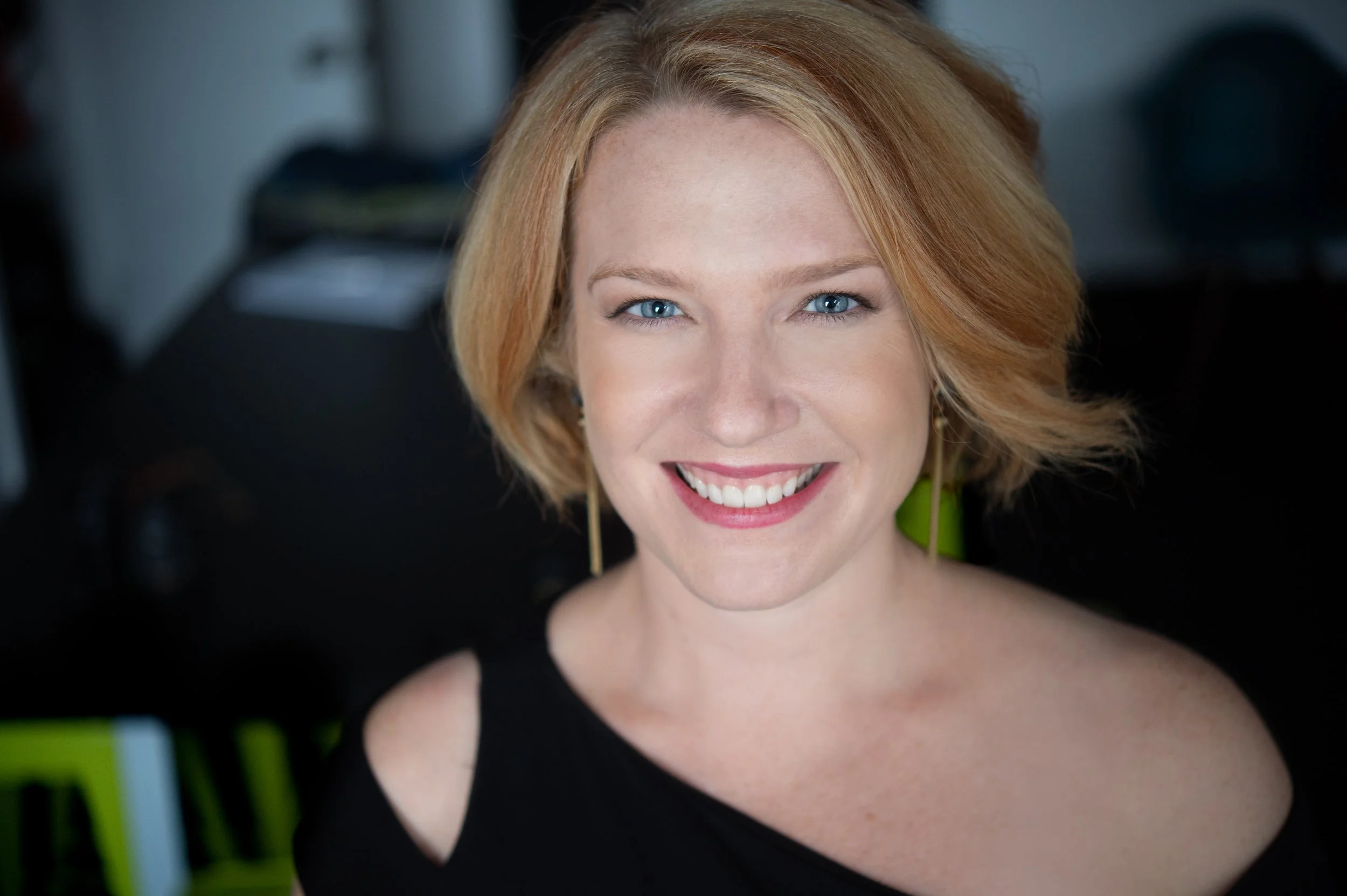 Close-up of a smiling woman with blue eyes and blonde hair, wearing a black dress and yellow earrings, indoors with a blurred background.