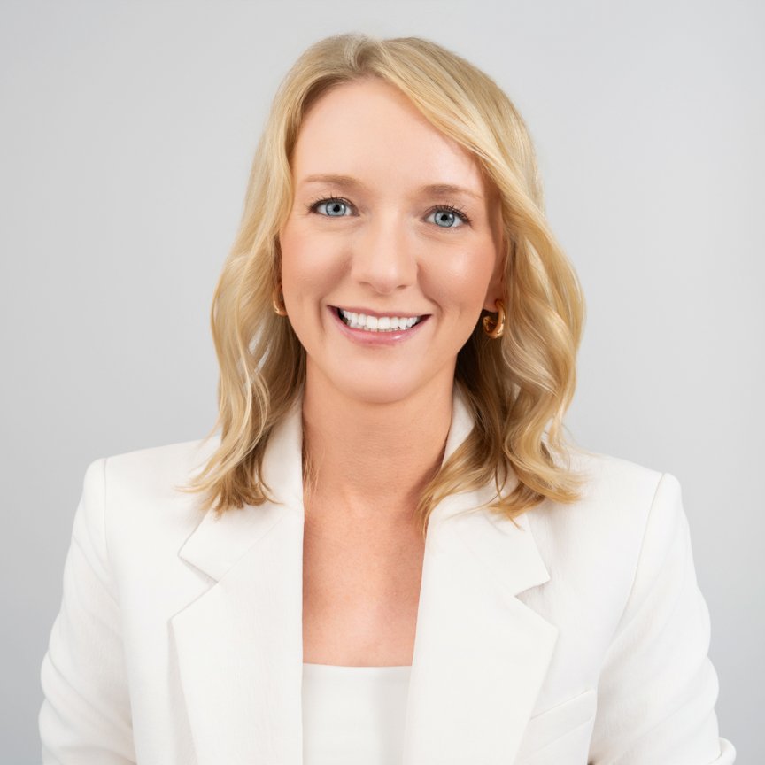 Portrait of a smiling woman with blonde hair, wearing a white blazer and gold earrings, against a plain light gray background.