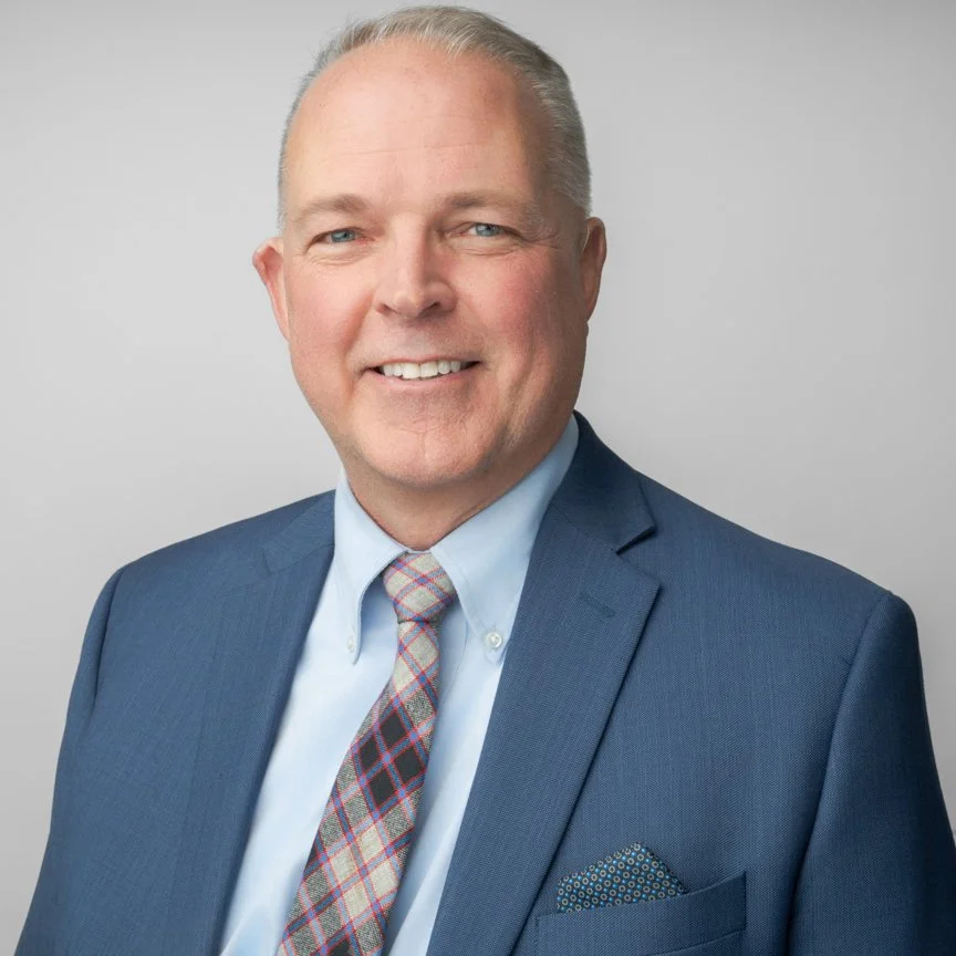 Professional headshot of a smiling middle-aged man in a blue suit, light blue shirt, patterned tie, and pocket square against a plain gray background.