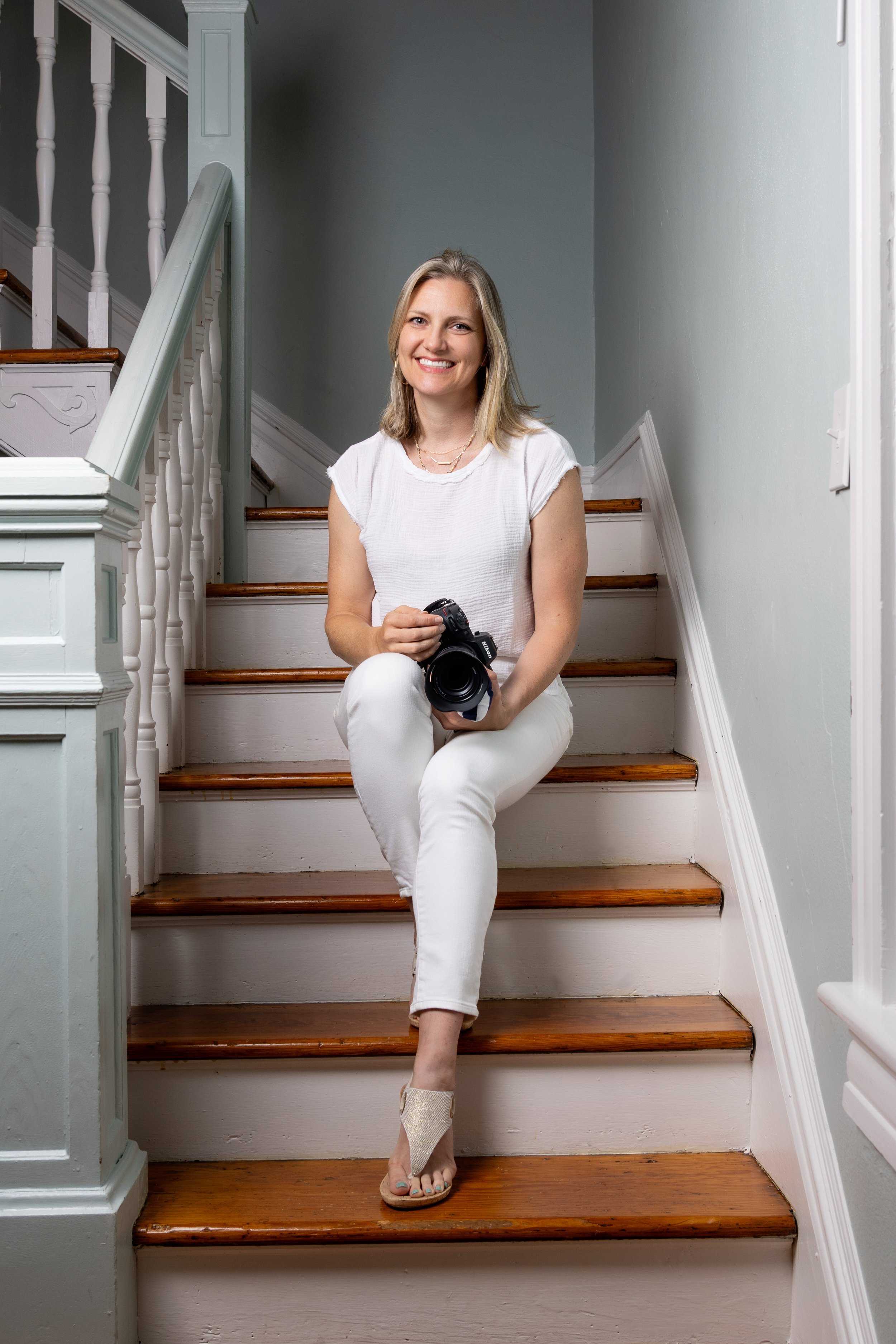 A woman with blonde hair sitting on wooden stairs, holding a camera and smiling.