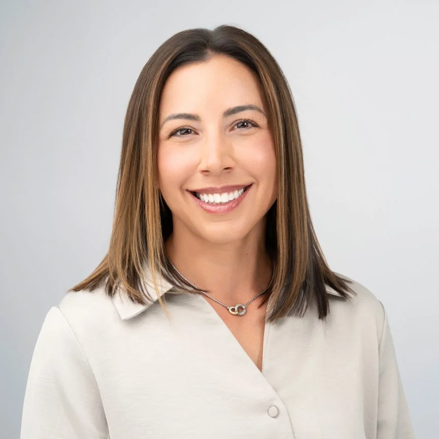 A smiling woman with straight brown hair, wearing a light-colored blouse and a silver necklace, against a plain gray background.