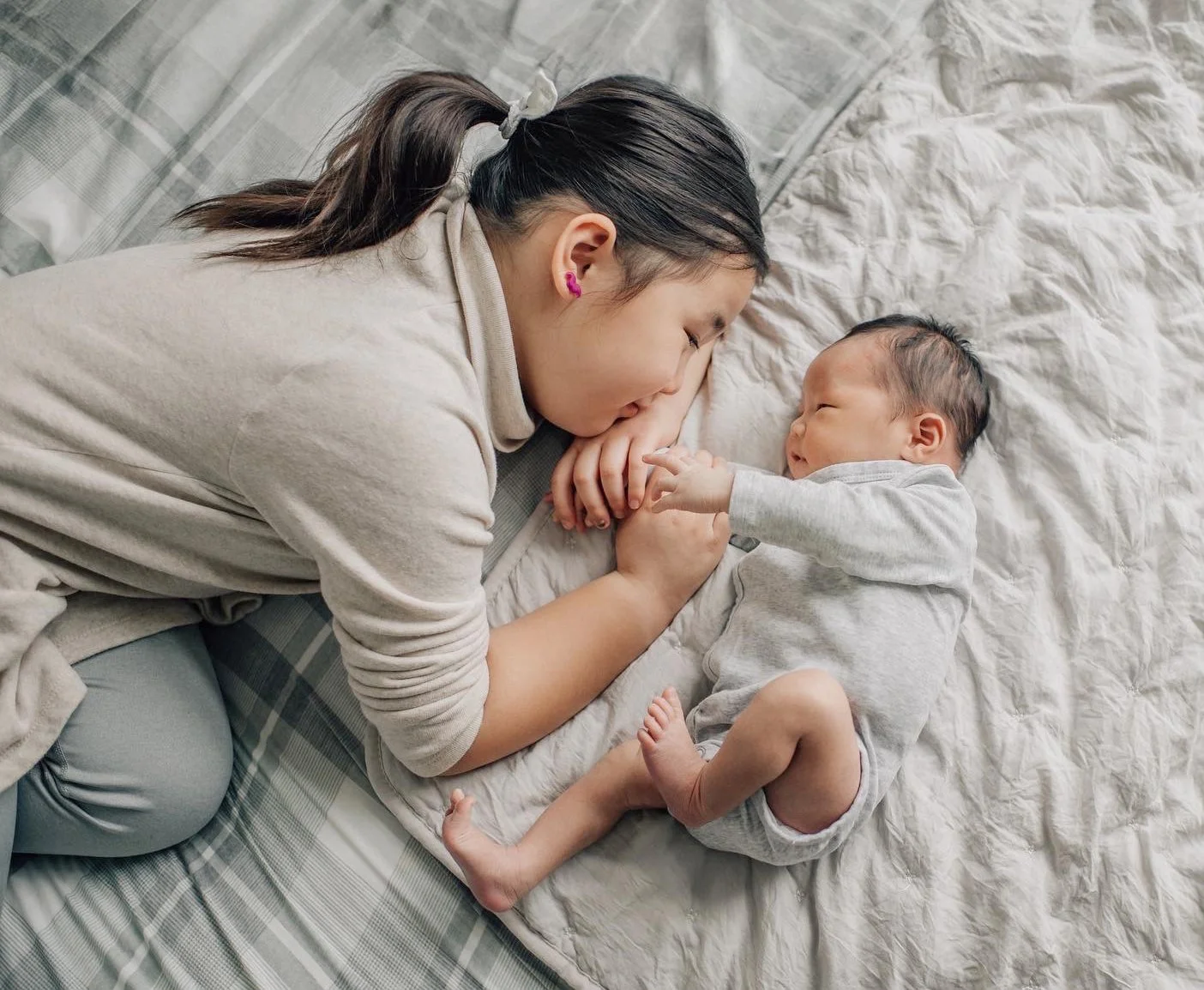 A woman and a baby lying on a bed, touching foreheads and holding hands. They are both wearing neutral-colored pajamas.