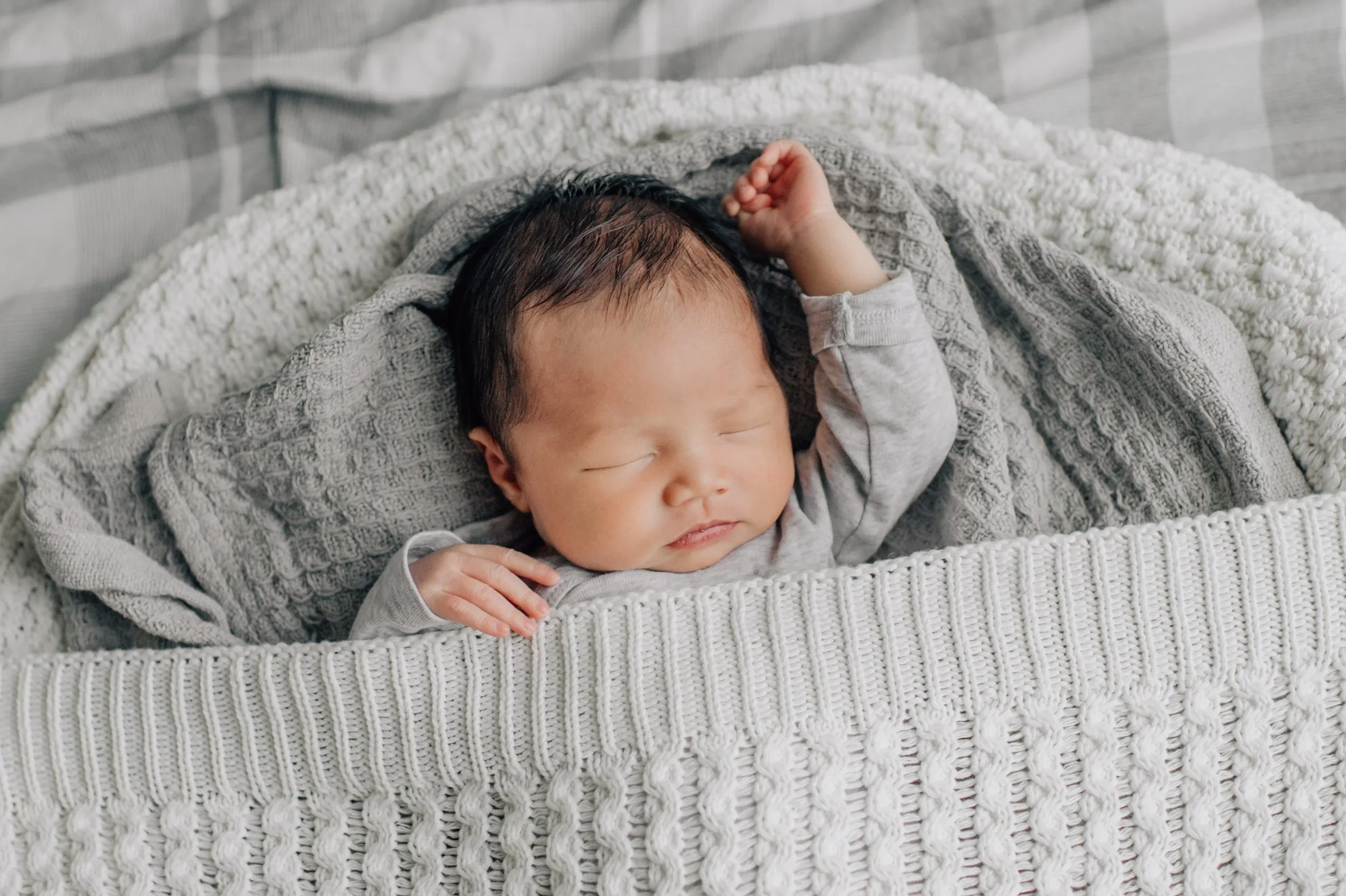 A sleeping baby with dark hair lying on a cream-colored knitted blanket in a crib.