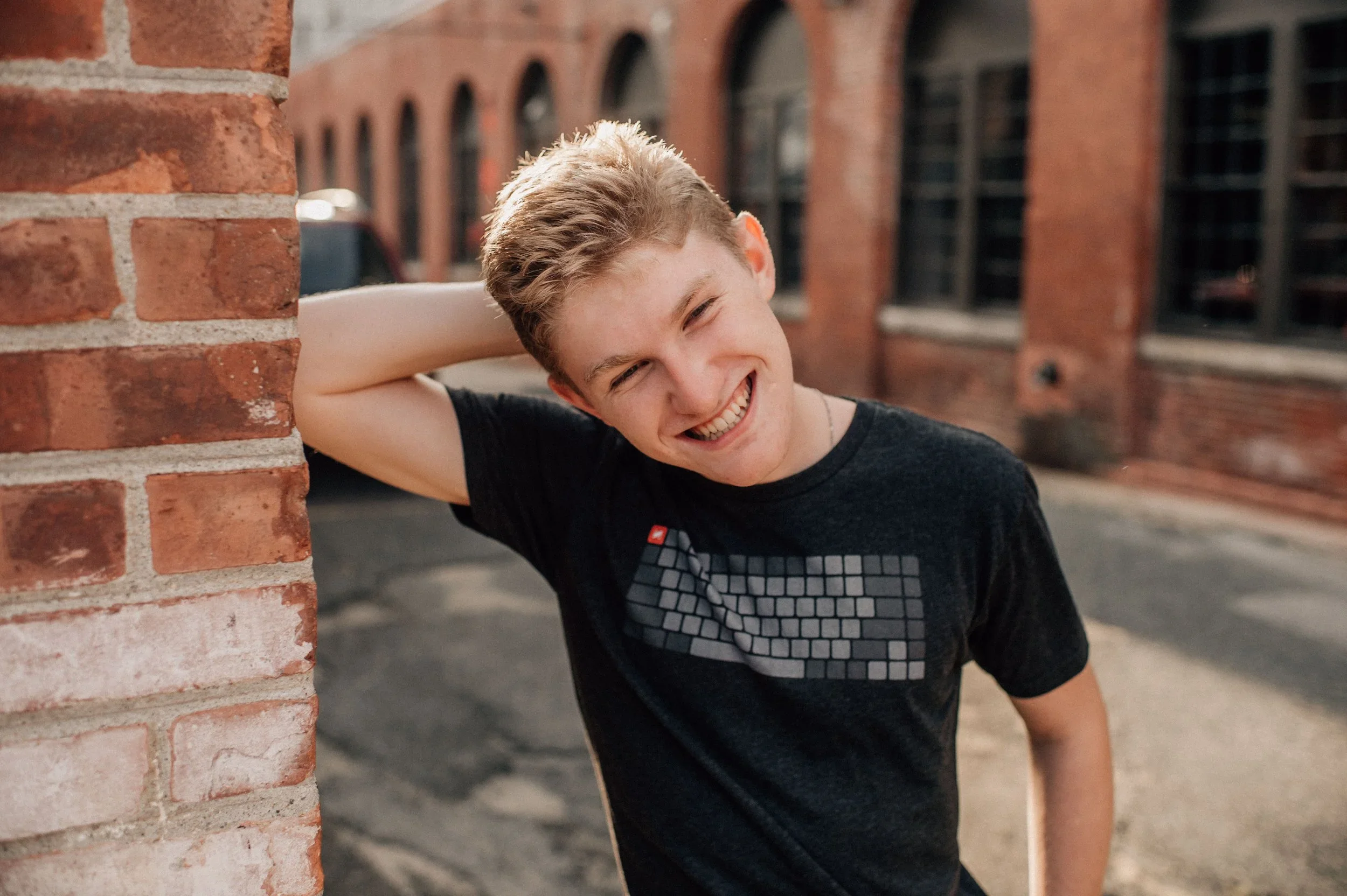 A young man with light brown hair smiling with his eyes closed, leaning against a brick wall outdoors.