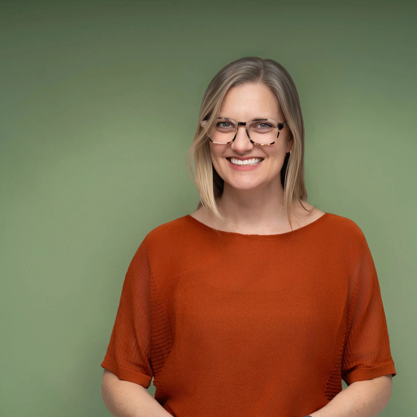 A woman with blonde hair, glasses, and a bright smile, wearing an orange top, standing against a green background.