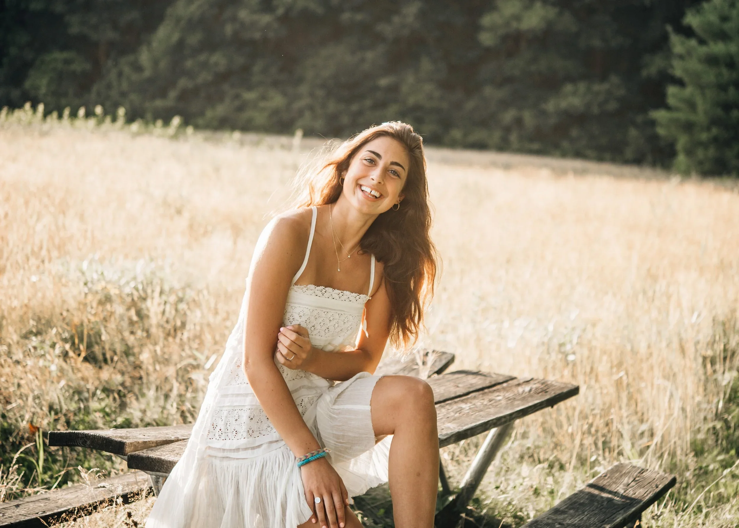 A young woman with long brown hair and a white dress sitting on a wooden bench outdoors in a field of tall grass with trees in the background, smiling at the camera.