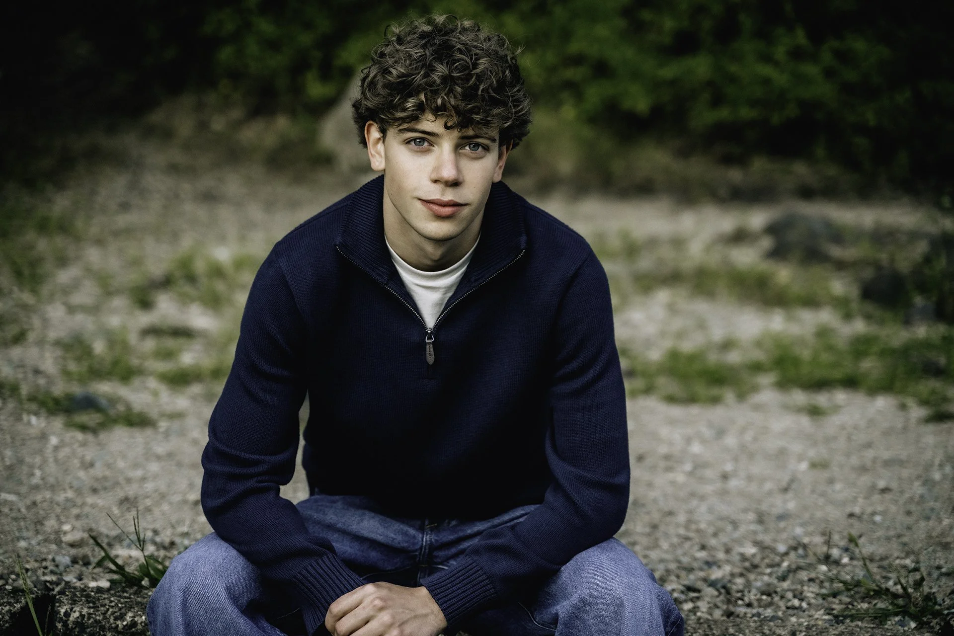 Young man with curly hair and blue eyes sitting outdoors on a dirt ground with greenery in the background.