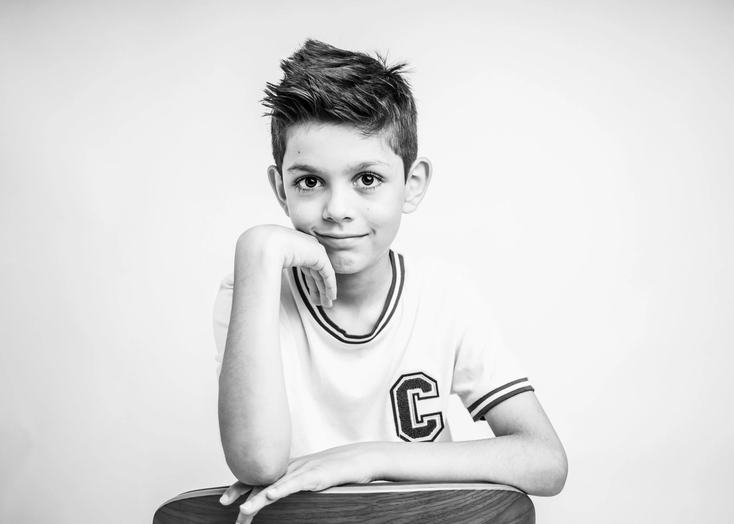 A young boy with short dark hair, wearing a white sports jersey with the letter "C" on it, sitting at a wooden table with his chin resting on his fist, smiling softly at the camera in a black and white portrait.