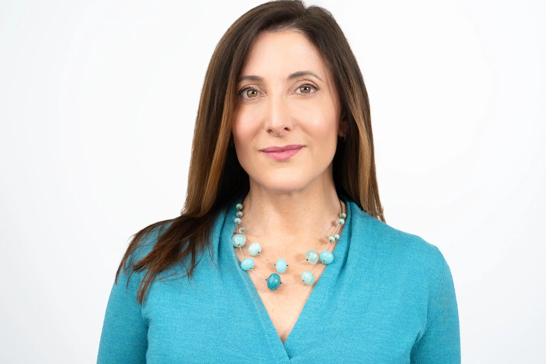 A woman with long brown hair wearing a turquoise top and a matching beaded necklace, standing against a plain white background.