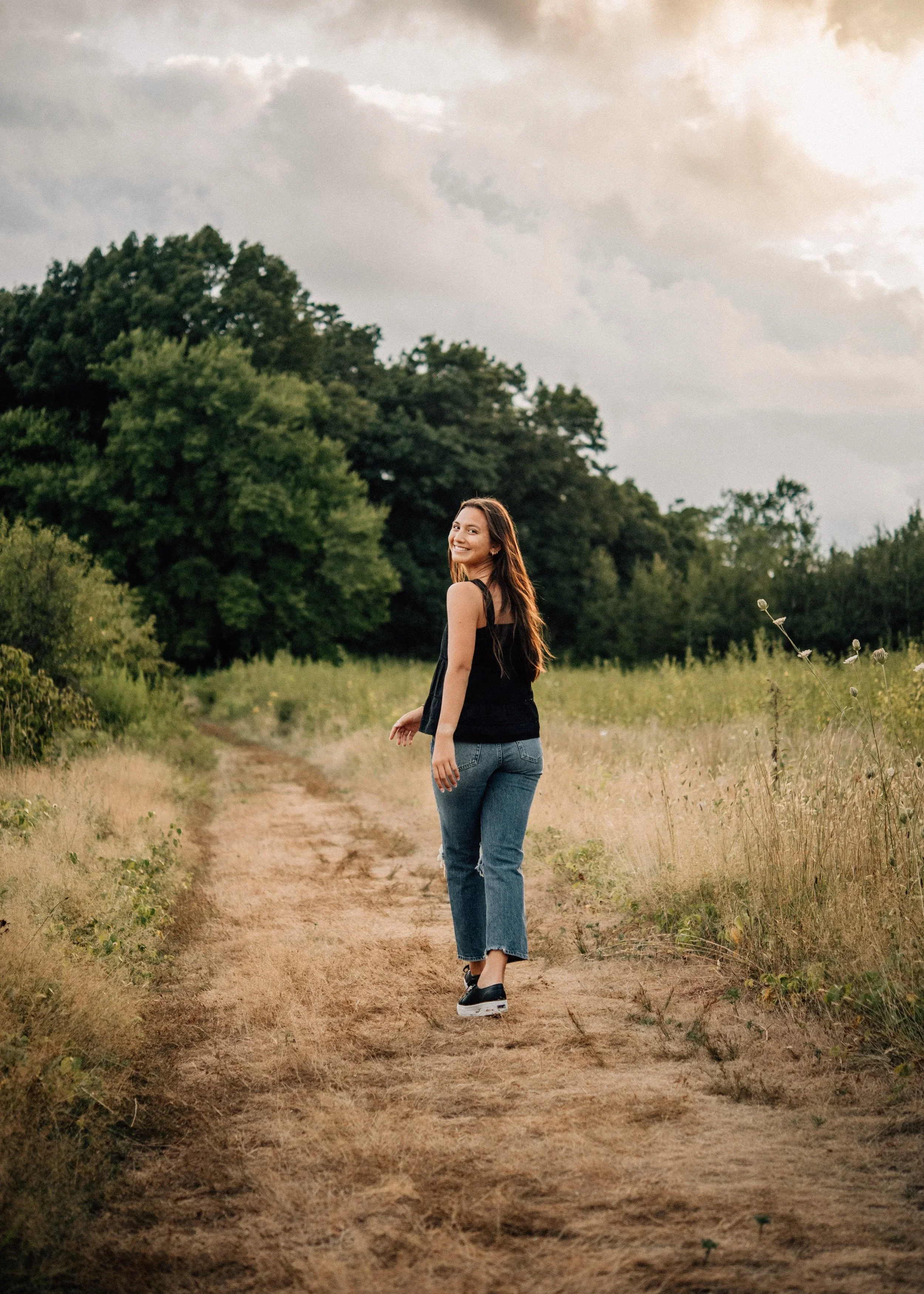 A smiling woman with long brown hair, wearing a black sleeveless top, jeans, and sneakers, walking on a dirt path in a grassy field with trees in the background under a cloudy sky.